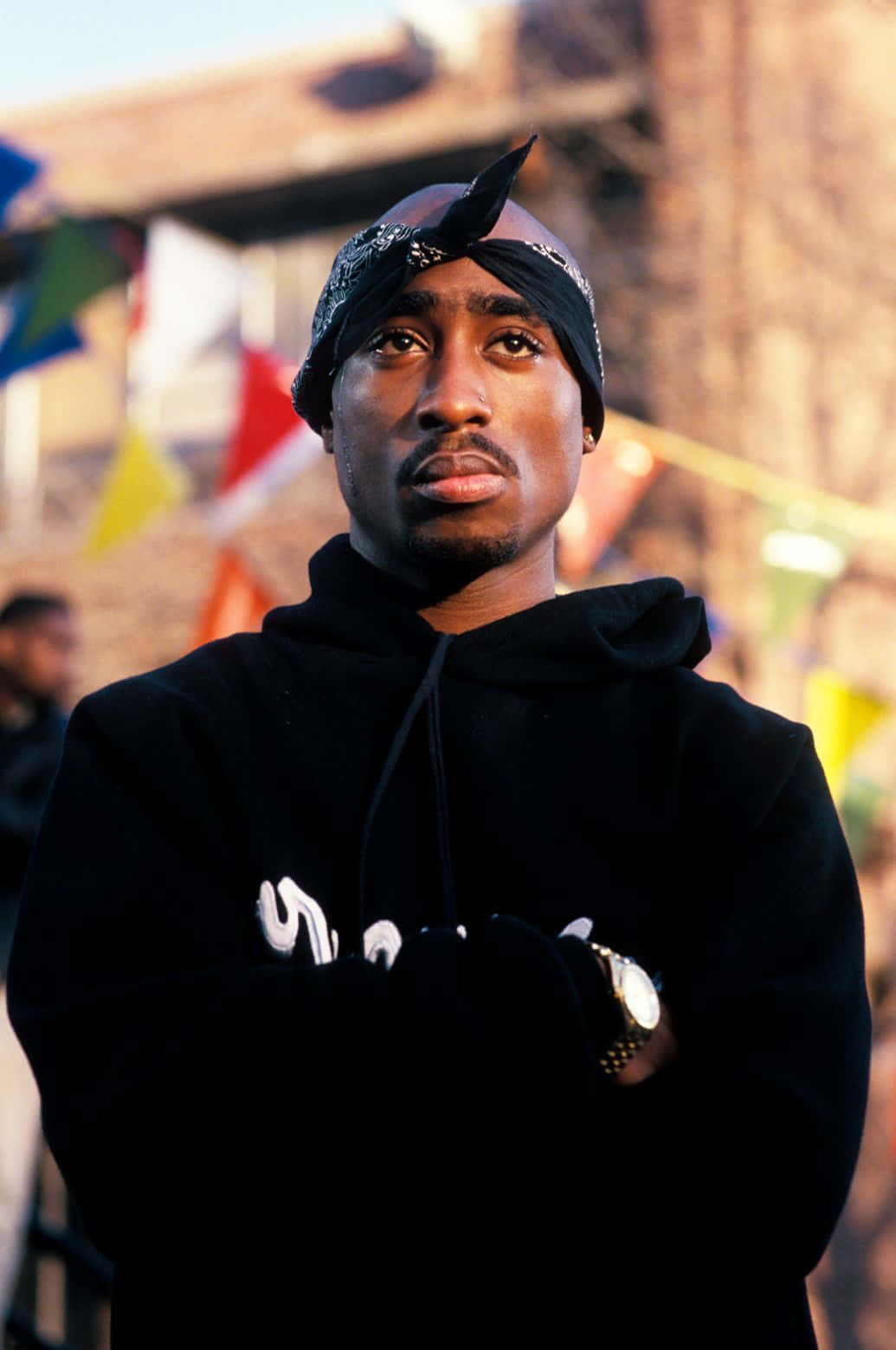 Tupac Shakur 身穿黑色連帽衫和頭巾，戶外站立，身後有色彩繽紛的旗幟。 — Tupac GIF 壁紙