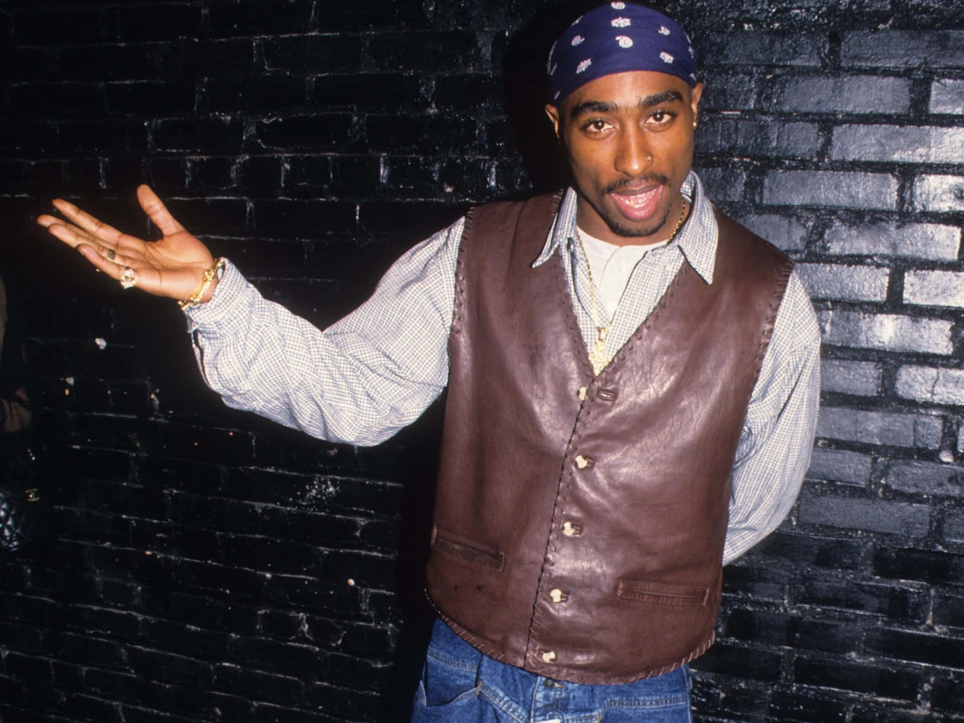 tupac 桌布 壁紙 — Tupac Shakur 自信地擺姿勢，背景是黑色磚牆，身穿棕色皮背心和藍色頭巾。