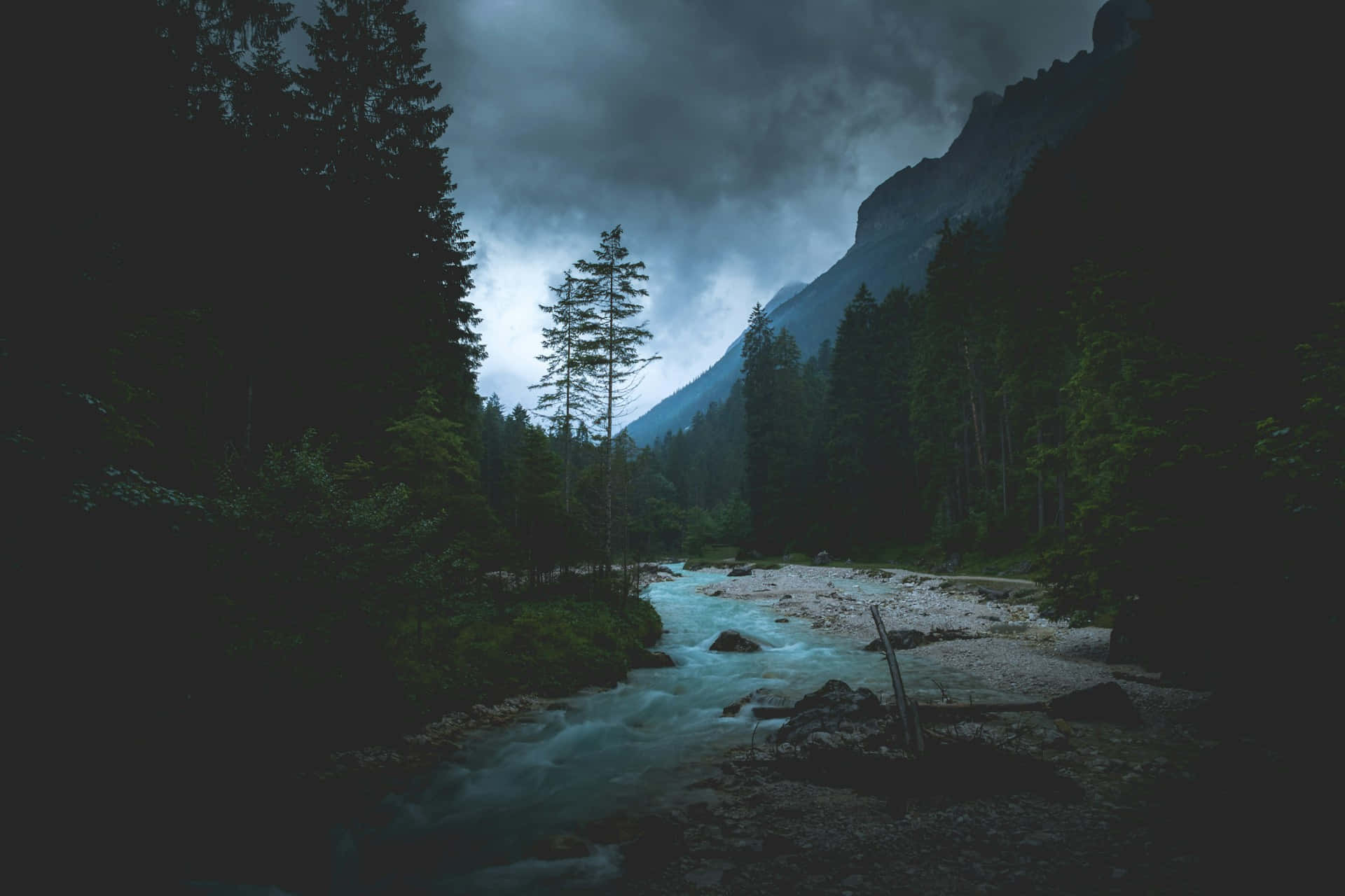  壁紙 — 一條湍急的河流流經黑暗的森林山谷，上方是暴風雨前的暮色天空。