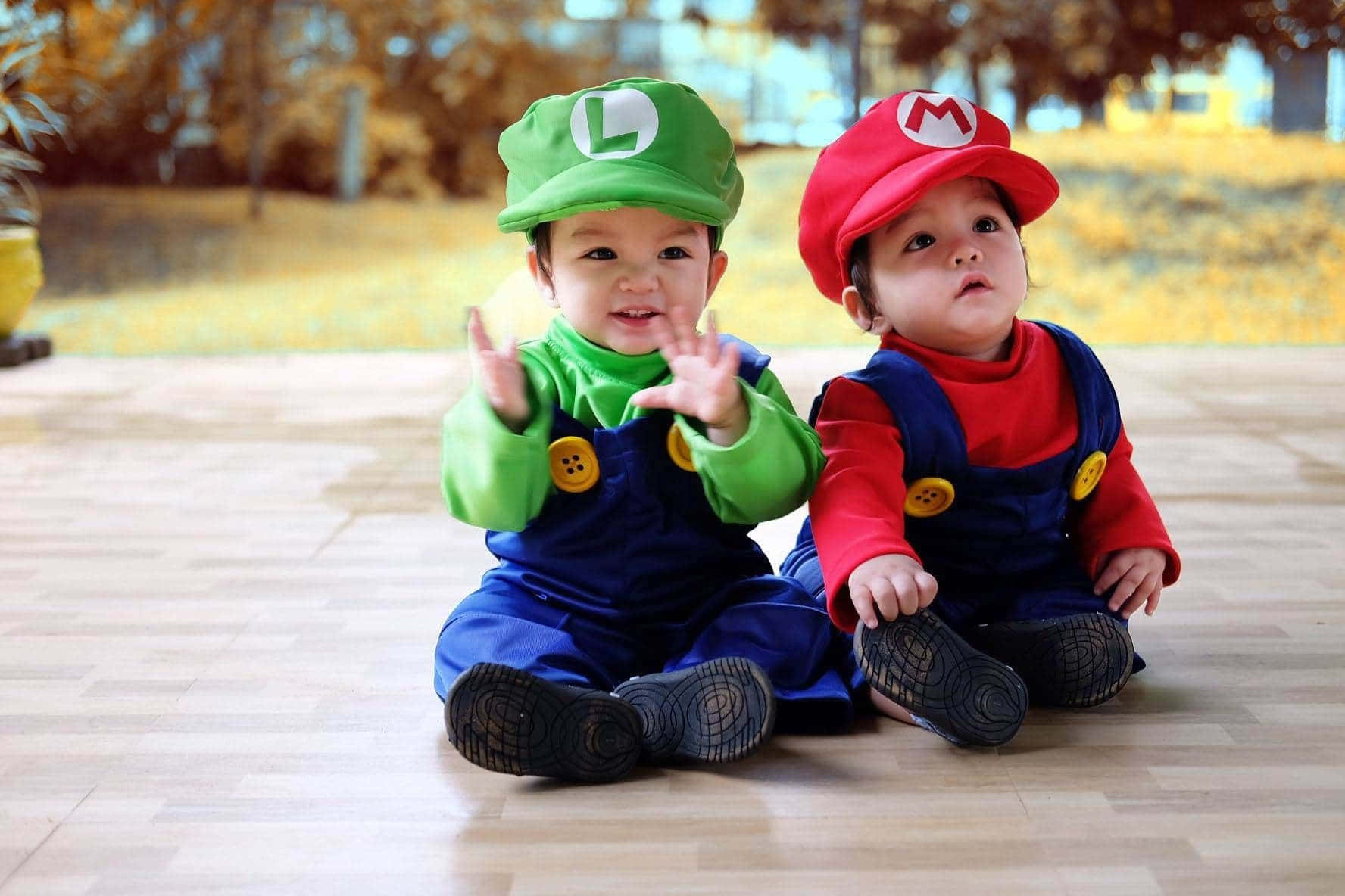 Baby Luigi 桌布 壁紙 — 兩個可愛的寶寶打扮成瑪利歐和路易吉，坐在木地板上開心地向鏡頭揮手。