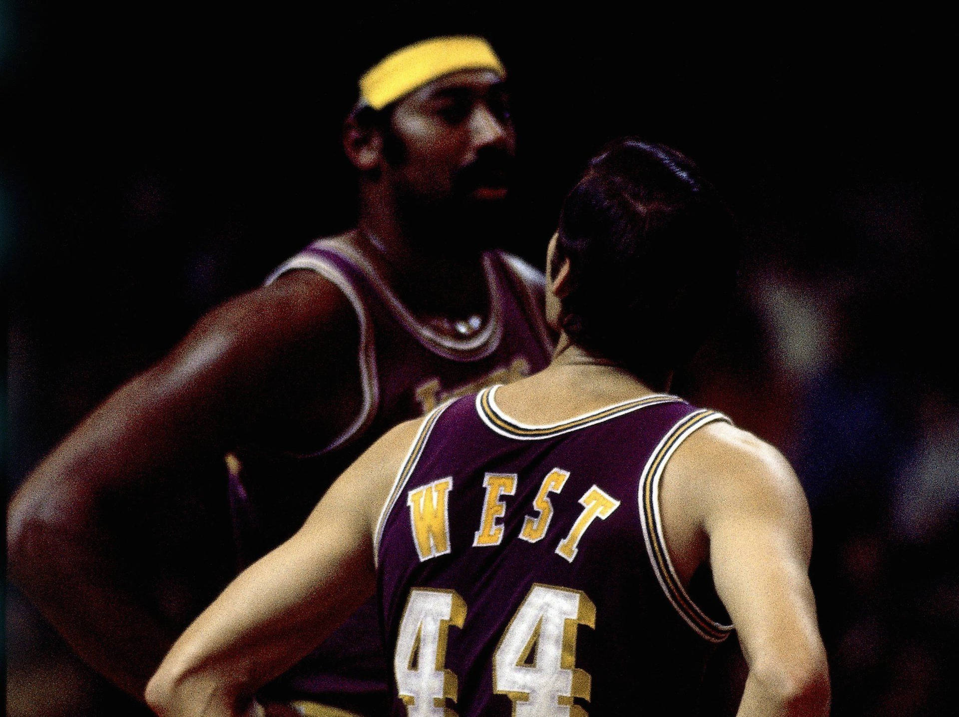 兩名身穿紫色和金色球衣的籃球運動員在場館燈光下激烈對峙。 — NBA 壁紙