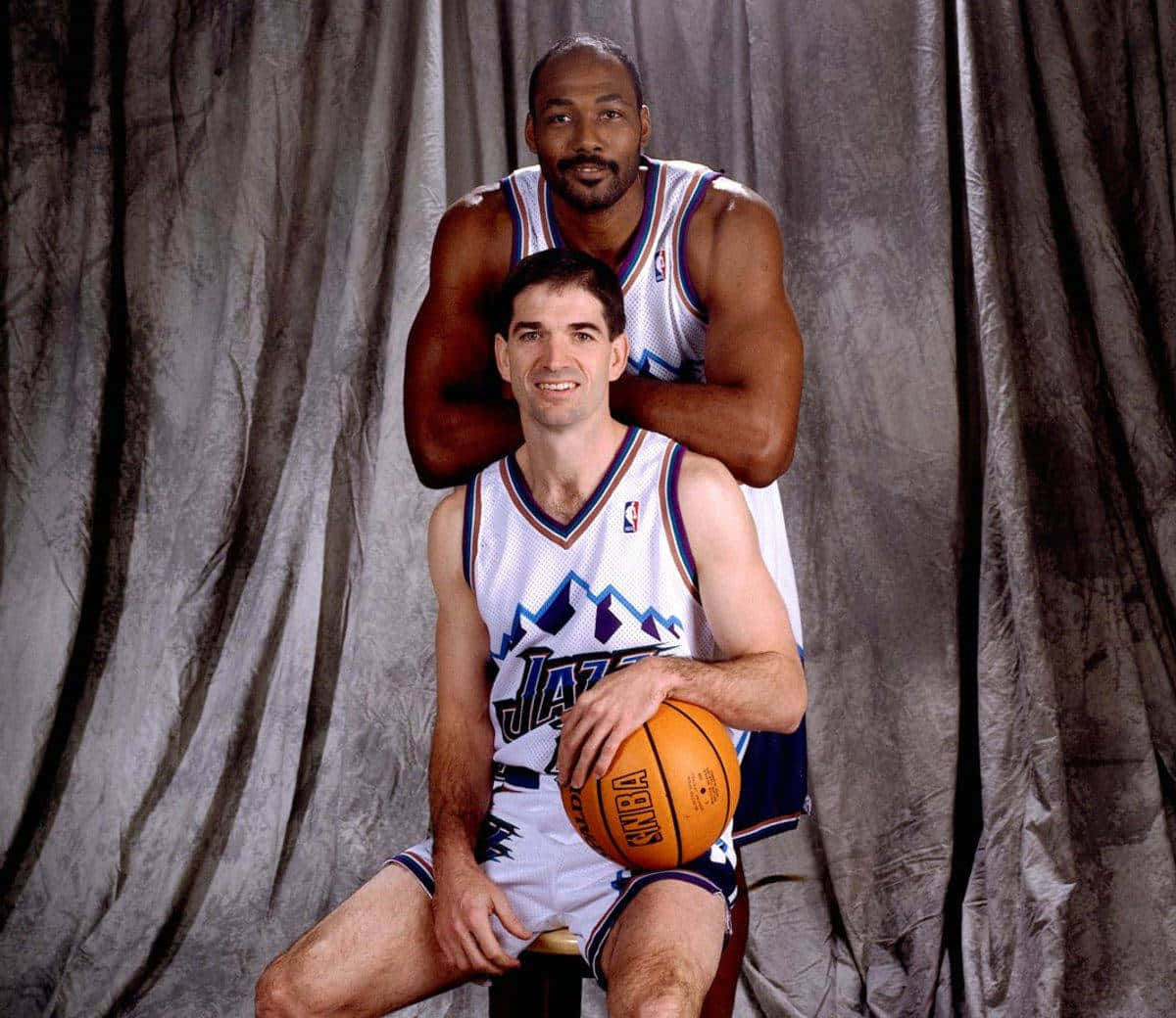 NBA 桌布 壁紙 — 兩名身穿猶他爵士隊球衣的籃球運動員擺姿勢，一人坐著持球，另一人站在他身後，背景為灰色窗簾。