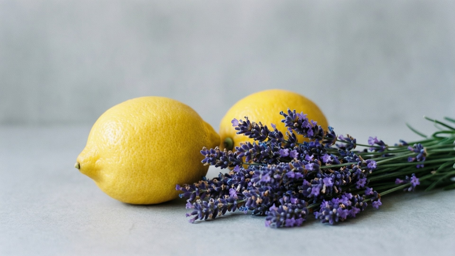 Lemon and Lavender Calm 壁紙 — 兩個鮮黃色的檸檬放在柔軟的灰色表面上，旁邊是一束紫色的薰衣草花。