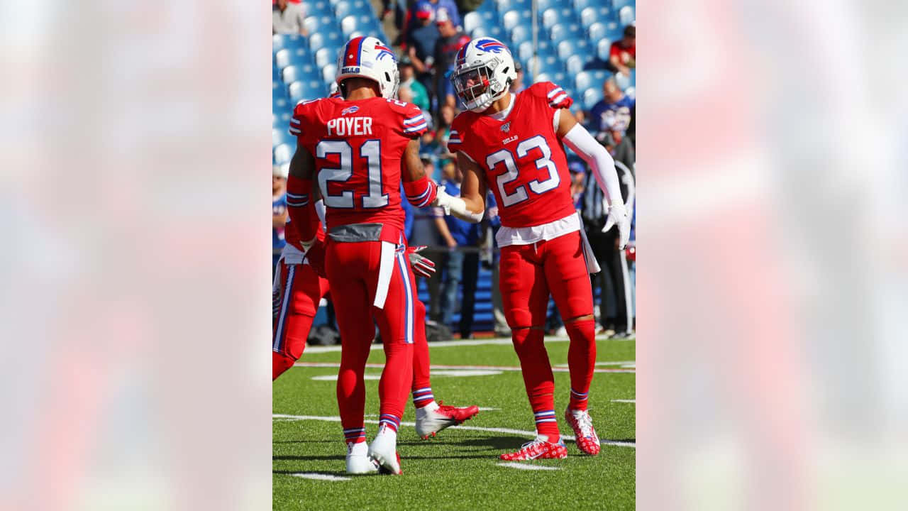 Jordan Poyer 桌布 壁紙 — 兩名身穿紅色球衣的布法羅比爾隊美式足球員在陽光明媚的綠色球場上擊掌，背景是體育場座位。
