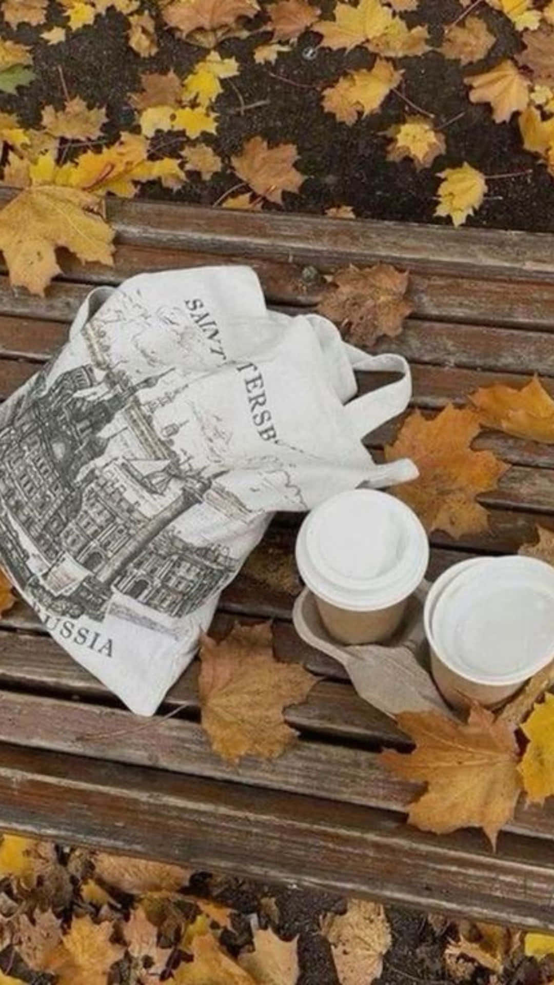  壁紙 — 兩個咖啡杯和一個手提袋放在一張木長椅上，周圍是秋天落下的金色楓葉。