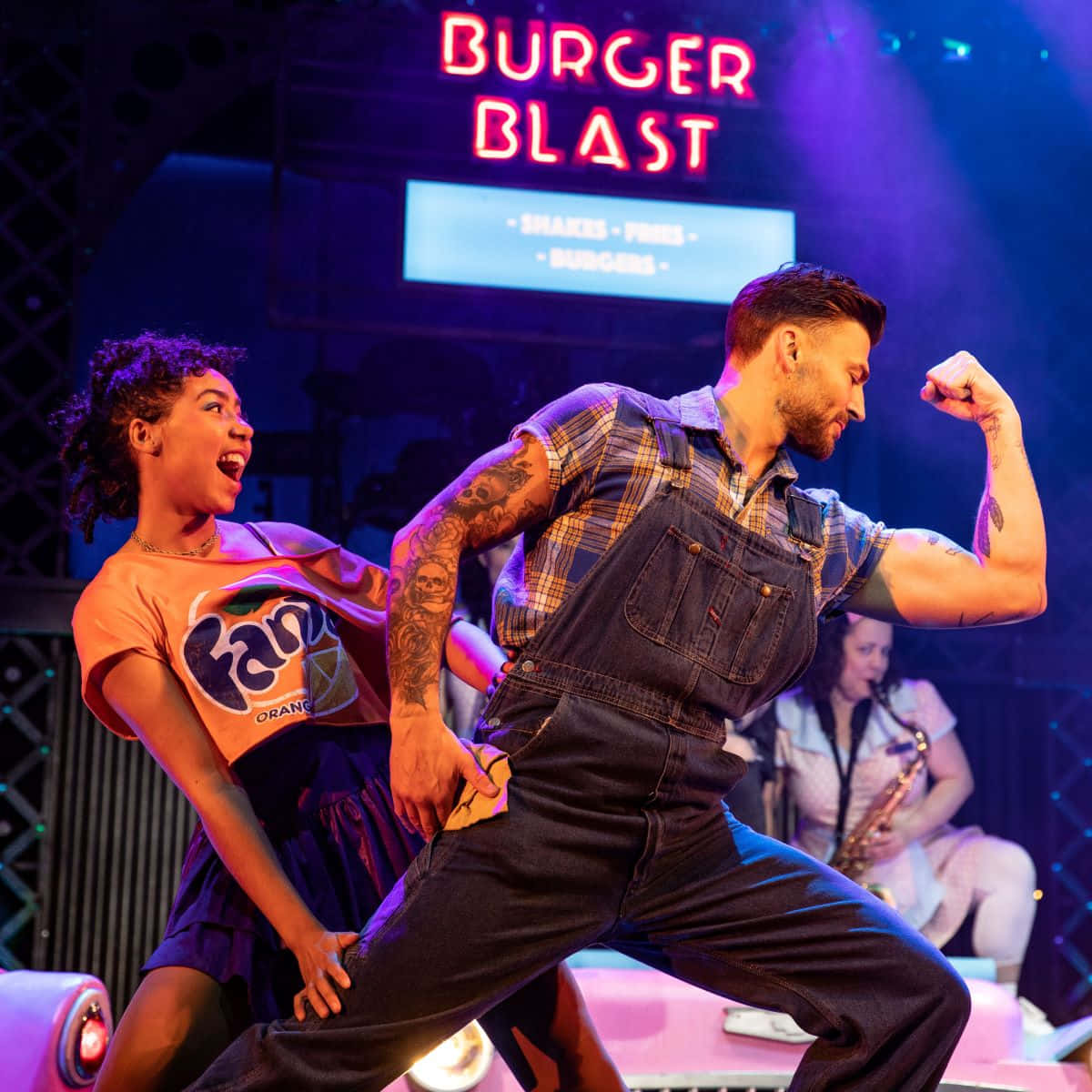 壁紙 — 兩位舞者在充滿活力的舞台燈光下，在發光的「Burger Blast」標誌下表演，背景有一位薩克斯風手。
