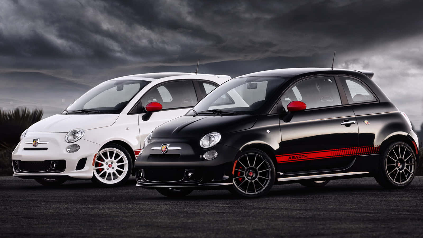 Fiat 500 桌布 壁紙 — 兩輛白色和黑色的 Fiat Abarth 汽車，帶有紅色點綴，在戲劇性的暴風雨天空下蓄勢待發。