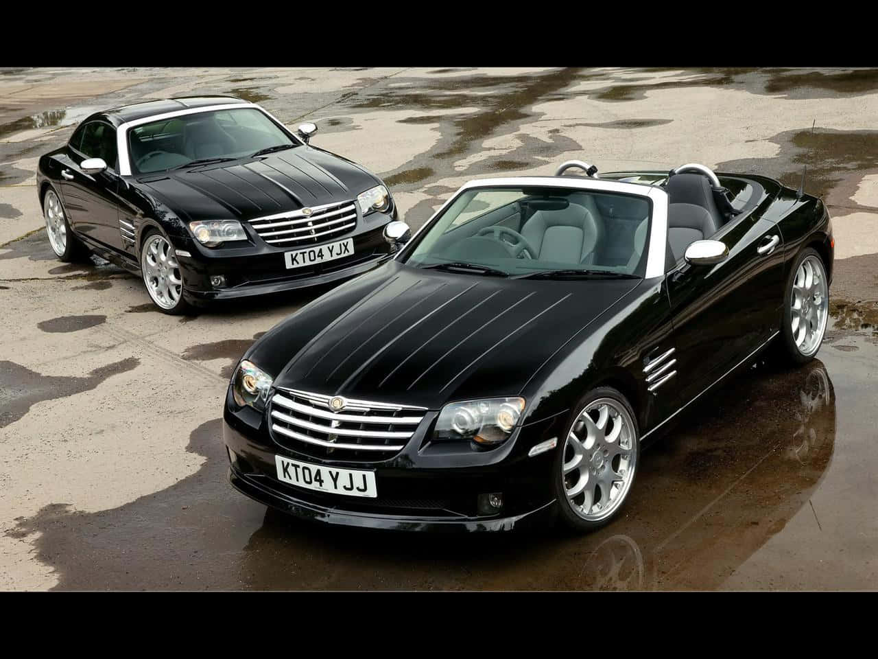 Chrysler Crossfire 桌布 壁紙 — 兩輛亮黑色克萊斯勒Crossfire跑車停在潮濕的路面上，反射出它們流暢的曲線和鉻合金輪圈。