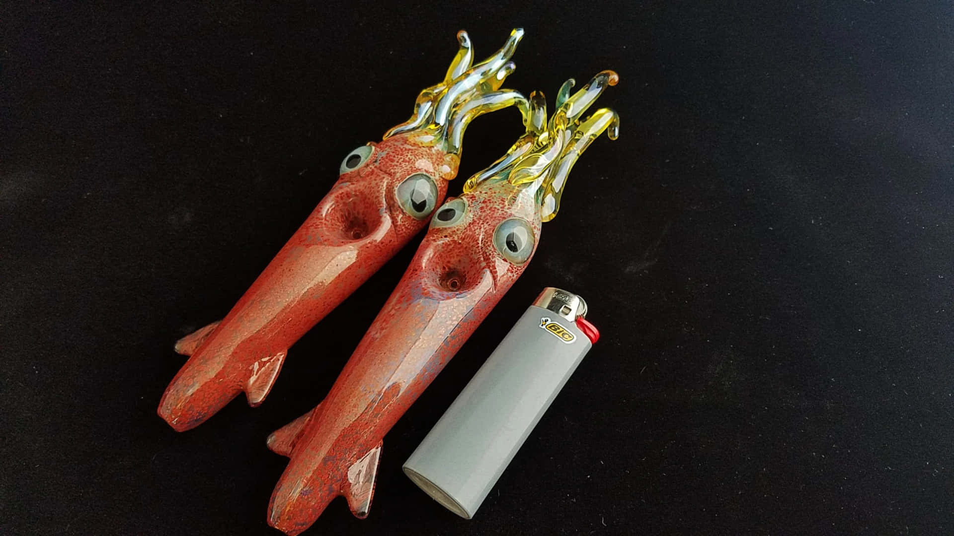  壁紙 — 兩個帶有玻璃眼睛和黃色觸手的亮面紅色玻璃魷魚雕塑，放在黑色表面上的一個銀色打火機旁邊。