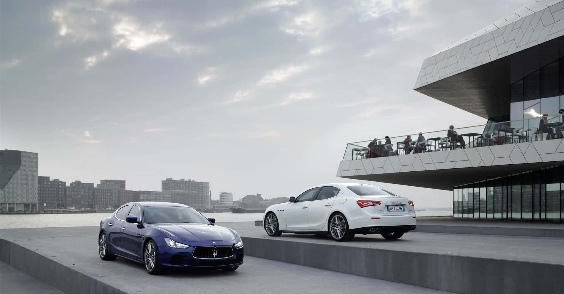 Maserati Ghibli 桌布 壁紙 — 兩輛豪華瑪莎拉蒂房車停放在現代海濱廣場上，背景是玻璃幕牆建築和城市天際線，天空多雲。