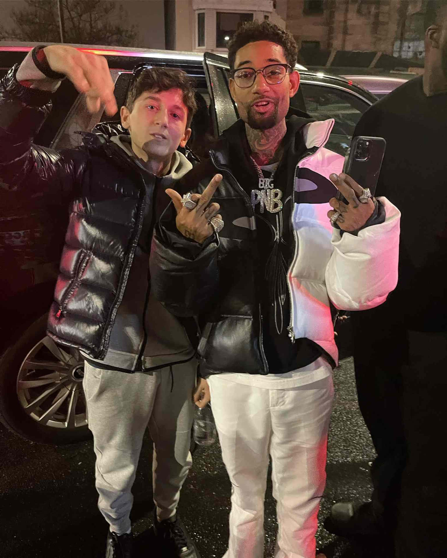  壁紙 — 兩名穿著羽絨外套的男子在夜間城市燈光下，於一輛汽車旁擺出各種手勢。