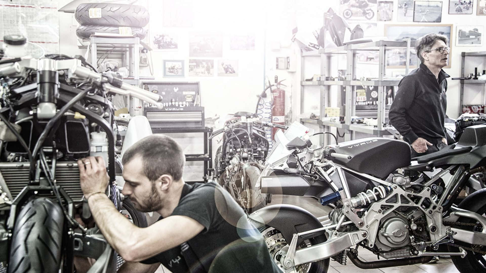 Vyrus Motorcycle 桌布 壁紙 — 兩名男子在一個雜亂的工作室中，處理訂製摩托車，工作室裡充滿了零件和復古海報。