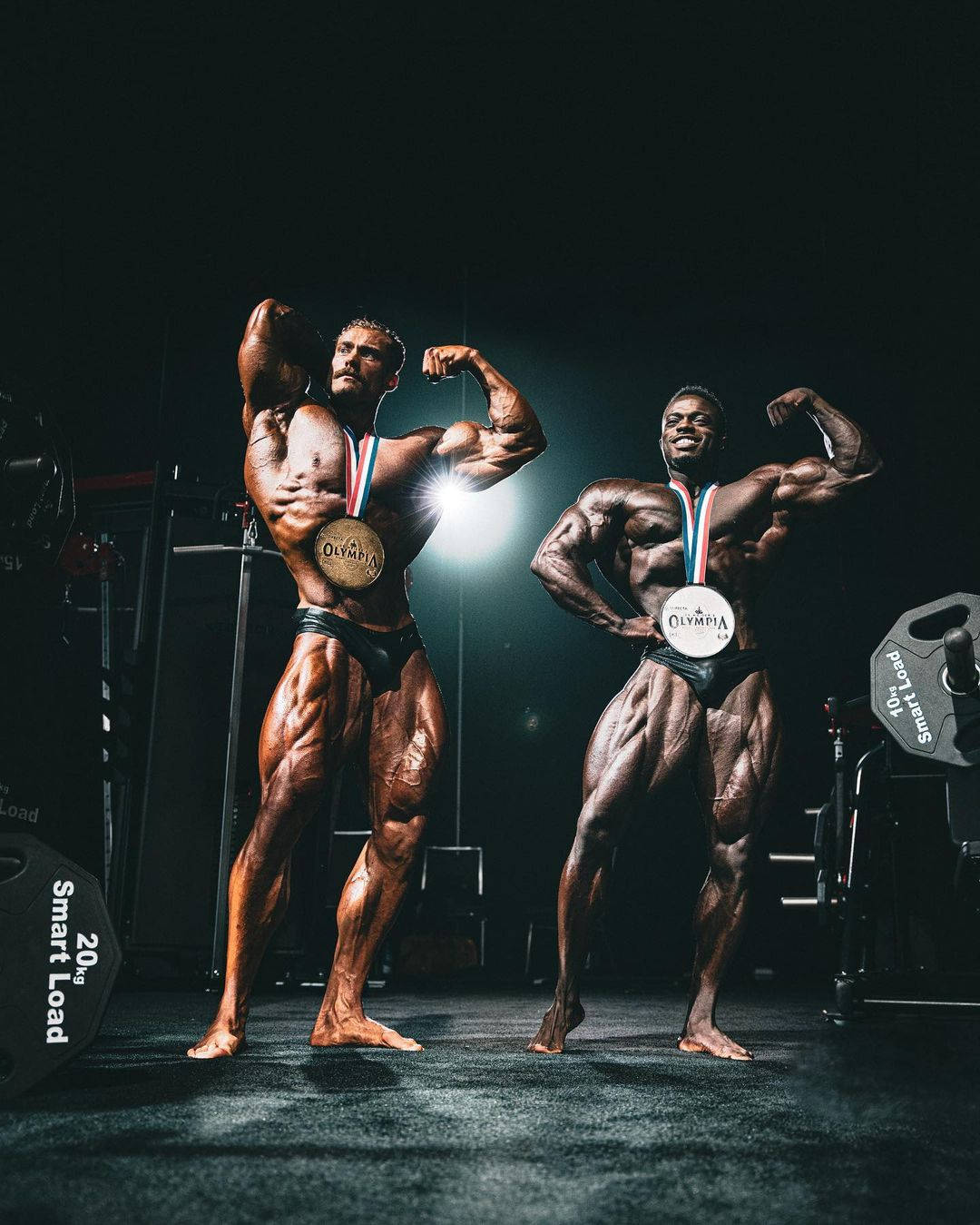兩位健美運動員在健身房聚光燈下拿著獎牌擺姿勢。 — Chris Bumstead 壁紙