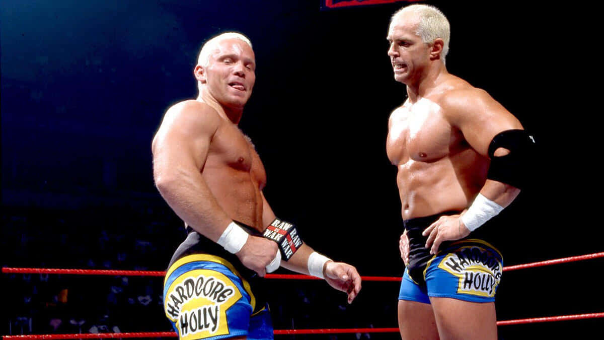 Crash Holly 桌布 壁紙 — 兩名肌肉發達的摔角手穿著藍色和黃色短褲，在一個光線充足、帶有紅色繩索的擂台上對峙。