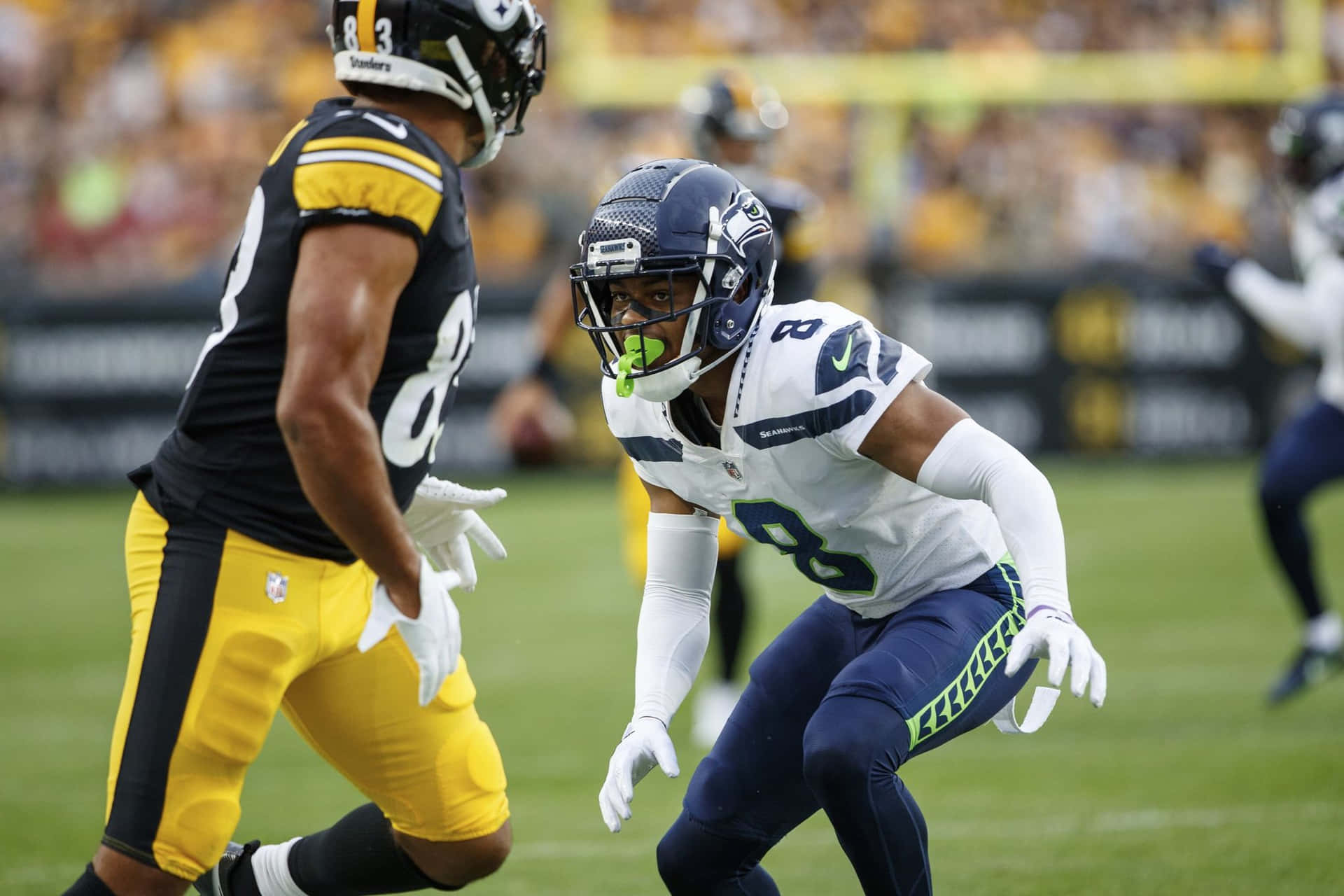  壁紙 — 兩位NFL球員在陽光明媚的球場上比賽，一位身穿Seahawks白色和藍色球衣，另一位身穿Steelers黑色和黃色球衣。