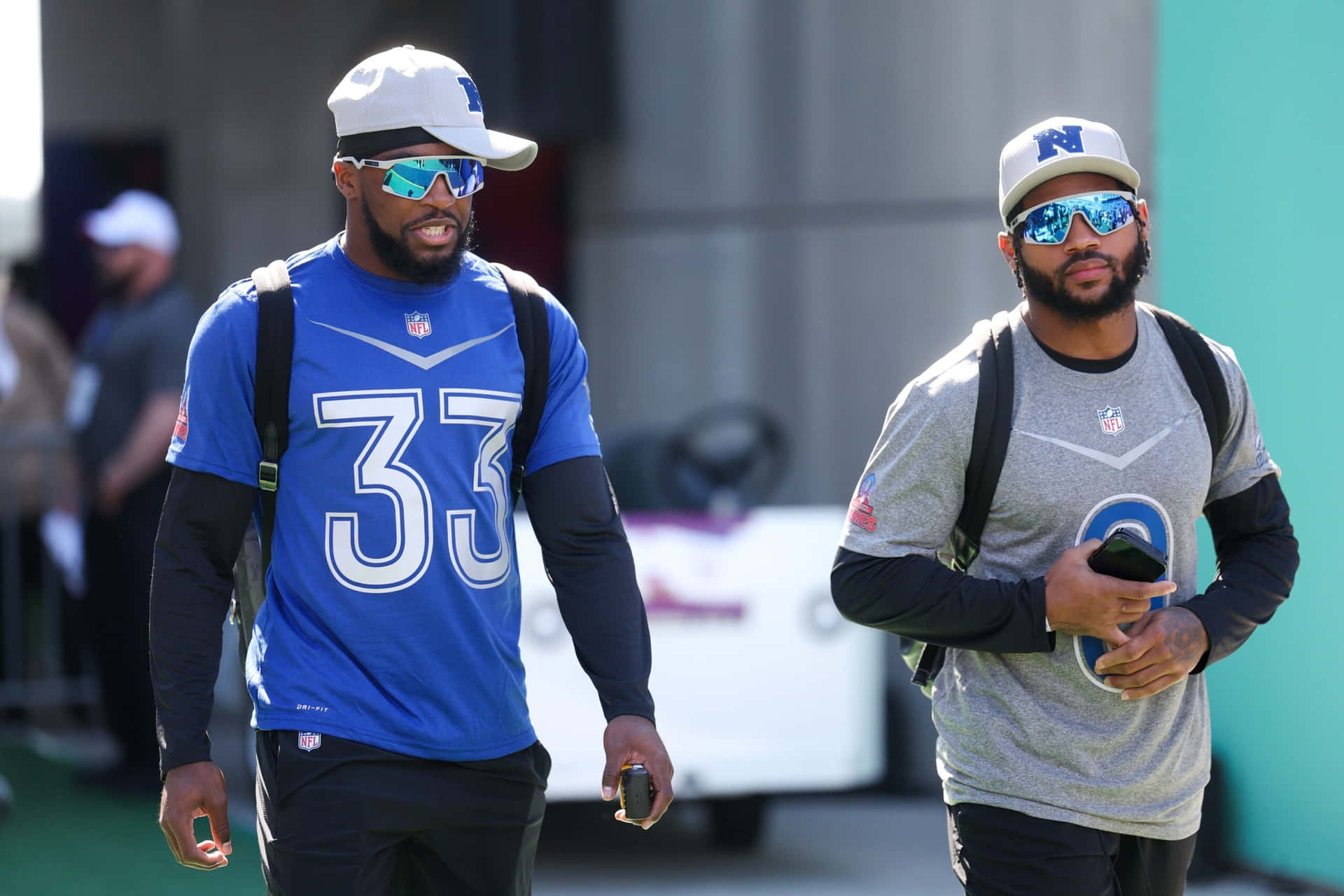  壁紙 — 兩名身穿球隊裝備的NFL球員戴著太陽眼鏡，手持電話在戶外行走。