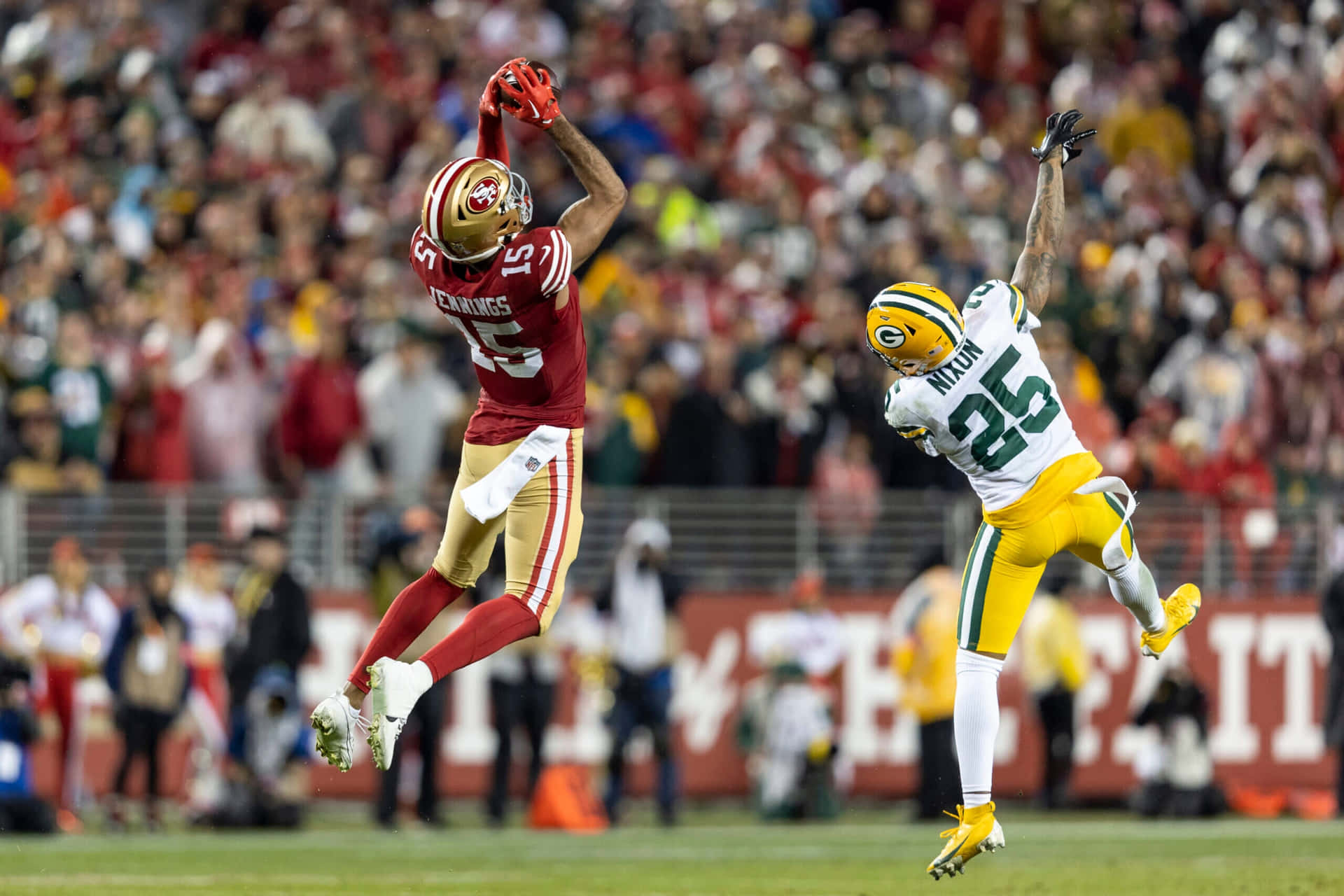  壁紙 — 兩位NFL球員在緊張的比賽時刻中跳起，一位身穿紅金色球衣伸手接球，另一位身穿綠黃色球衣防守。