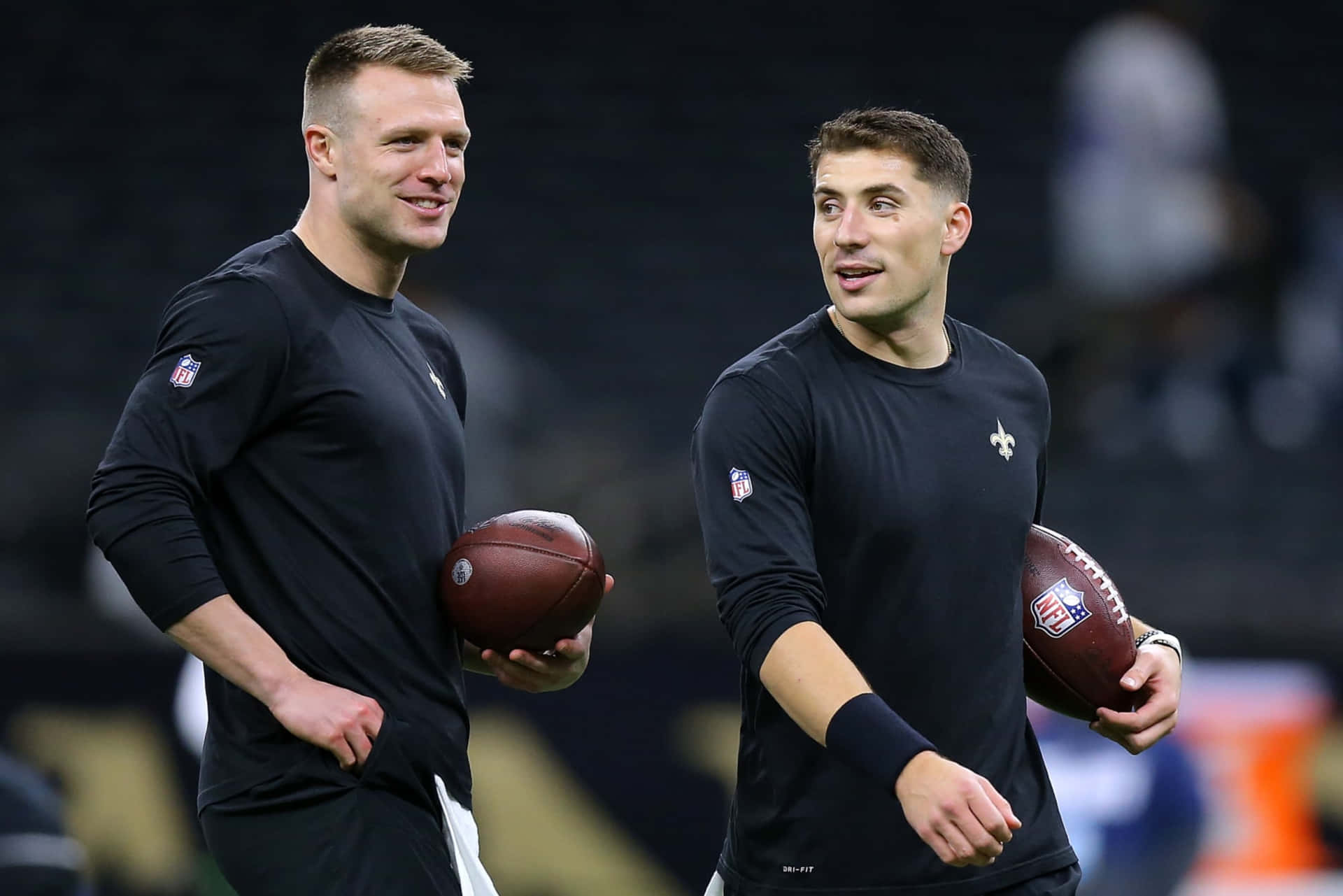  壁紙 — 兩位身穿黑色訓練裝備的NFL四分衛並肩而行，在昏暗的球場上手持橄欖球。