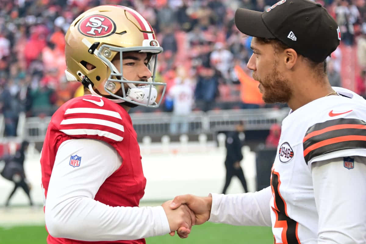  壁紙 — 兩位NFL四分衛在場上握手，一人身穿紅金色裝備，另一人身穿白棕色裝備，背後體育館人群模糊。