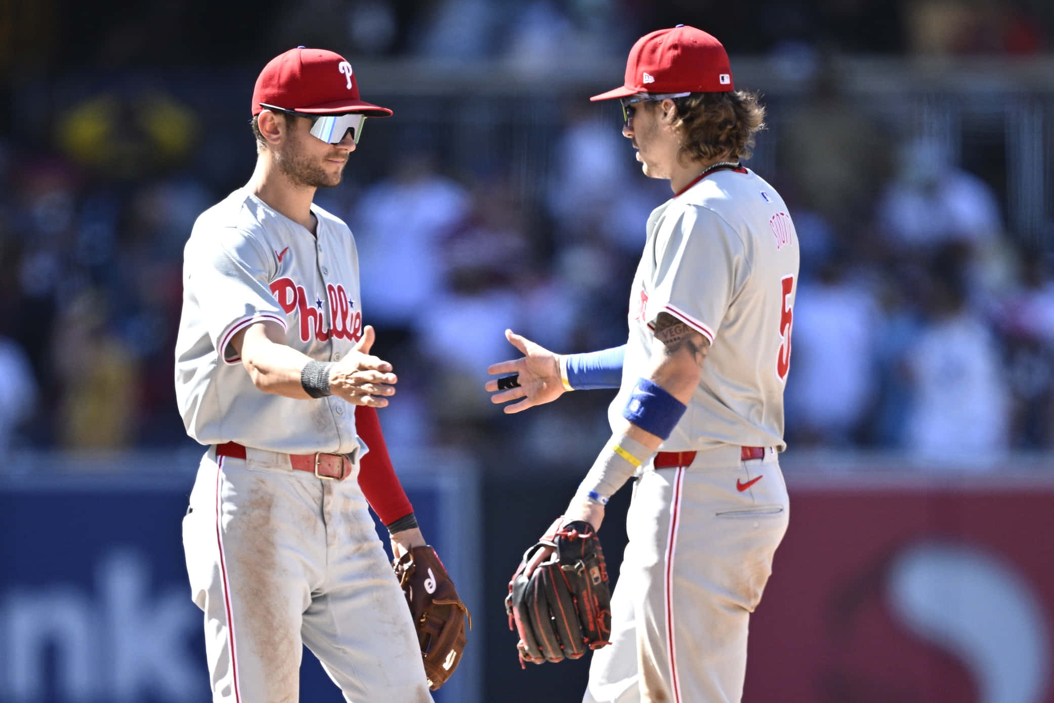 Trea Turner Phillies 桌布 壁紙 — 兩名身穿白色球衣的費城費城人棒球選手在陽光明媚的球場上握手，背景有模糊的觀眾。