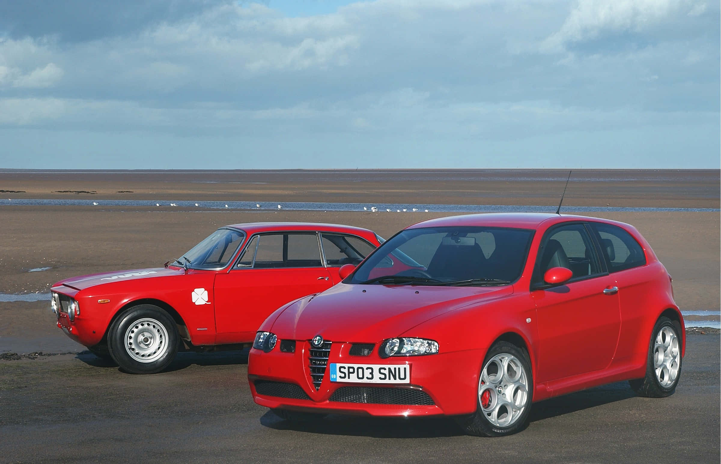 Alfa Romeo 147 桌布 壁紙 — 兩輛紅色愛快羅密歐汽車停泊在濕潤的潮灘上，天空多雲，遠處有鳥兒。