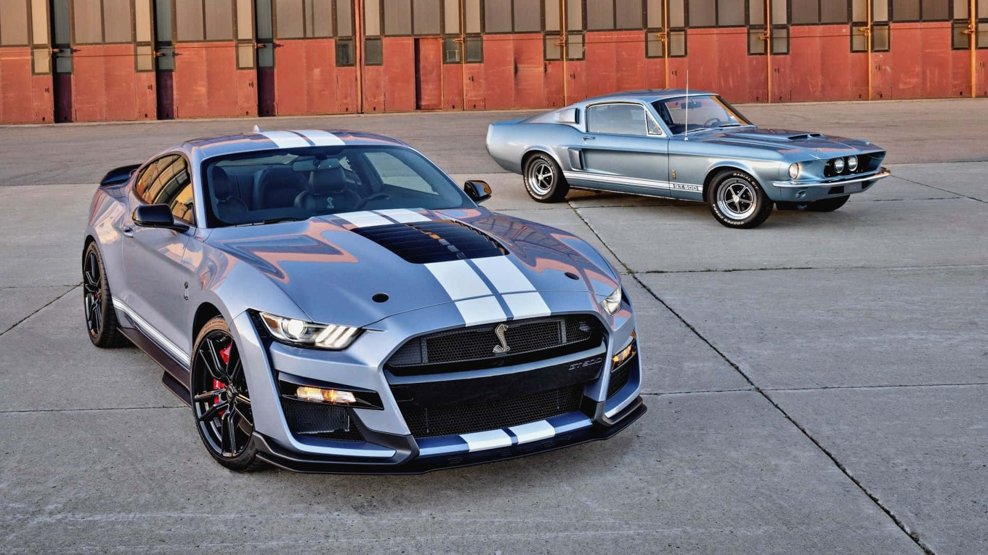  壁紙 — 兩輛帶有白色賽車條紋的銀色Shelby Mustang停放在紅磚建築前的混凝土上。