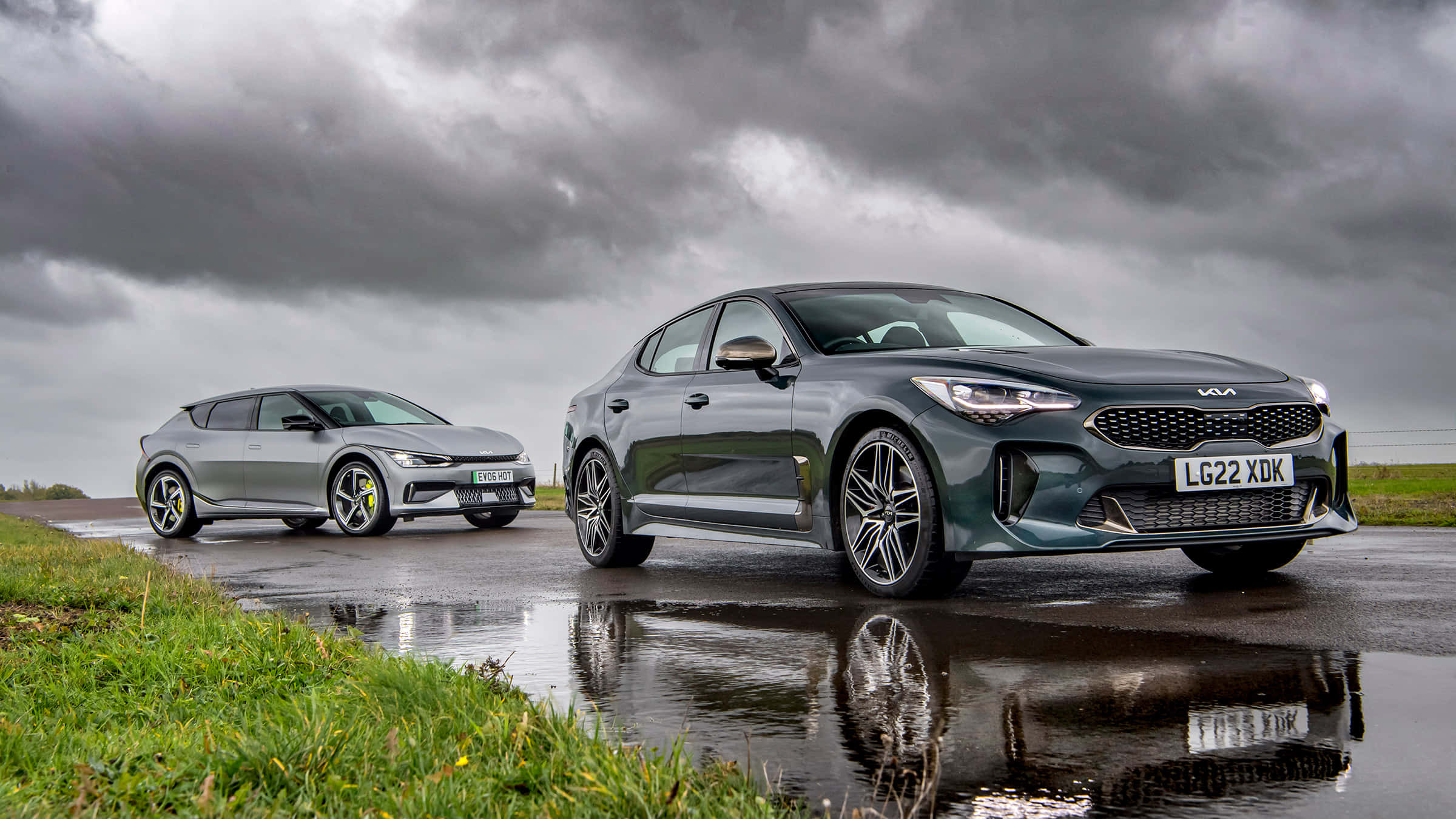 Kia Stinger 桌布 壁紙 — 兩輛時尚的Kia電動車在潮濕的道路上閃閃發光，背景是戲劇性的暴風雨天空，並映照著草地的邊緣。
