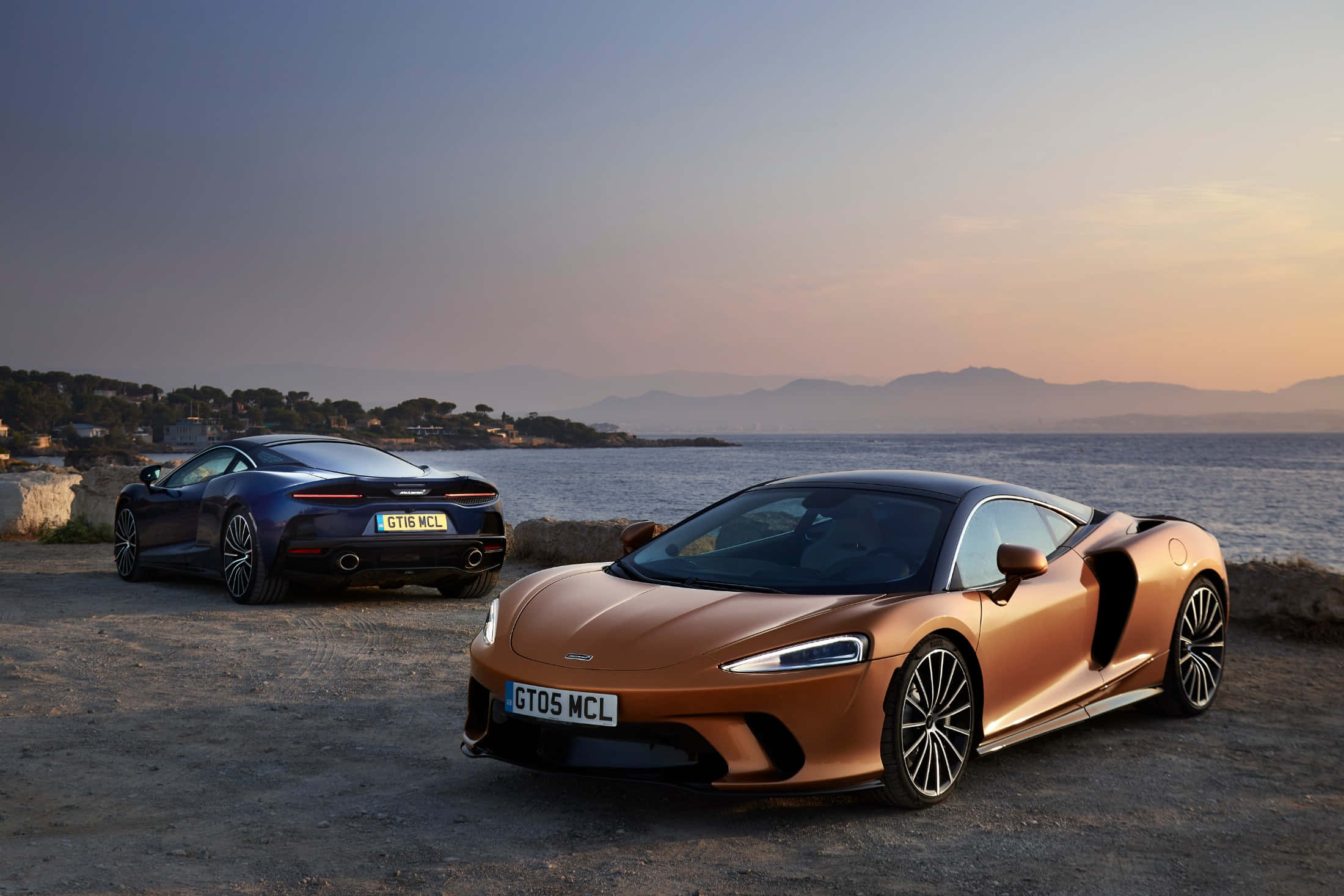 Mclaren Gt 桌布 壁紙 — 兩輛時尚的McLaren跑車在日落色調中閃耀，停泊在俯瞰平靜水域和遠山的海岸懸崖旁。