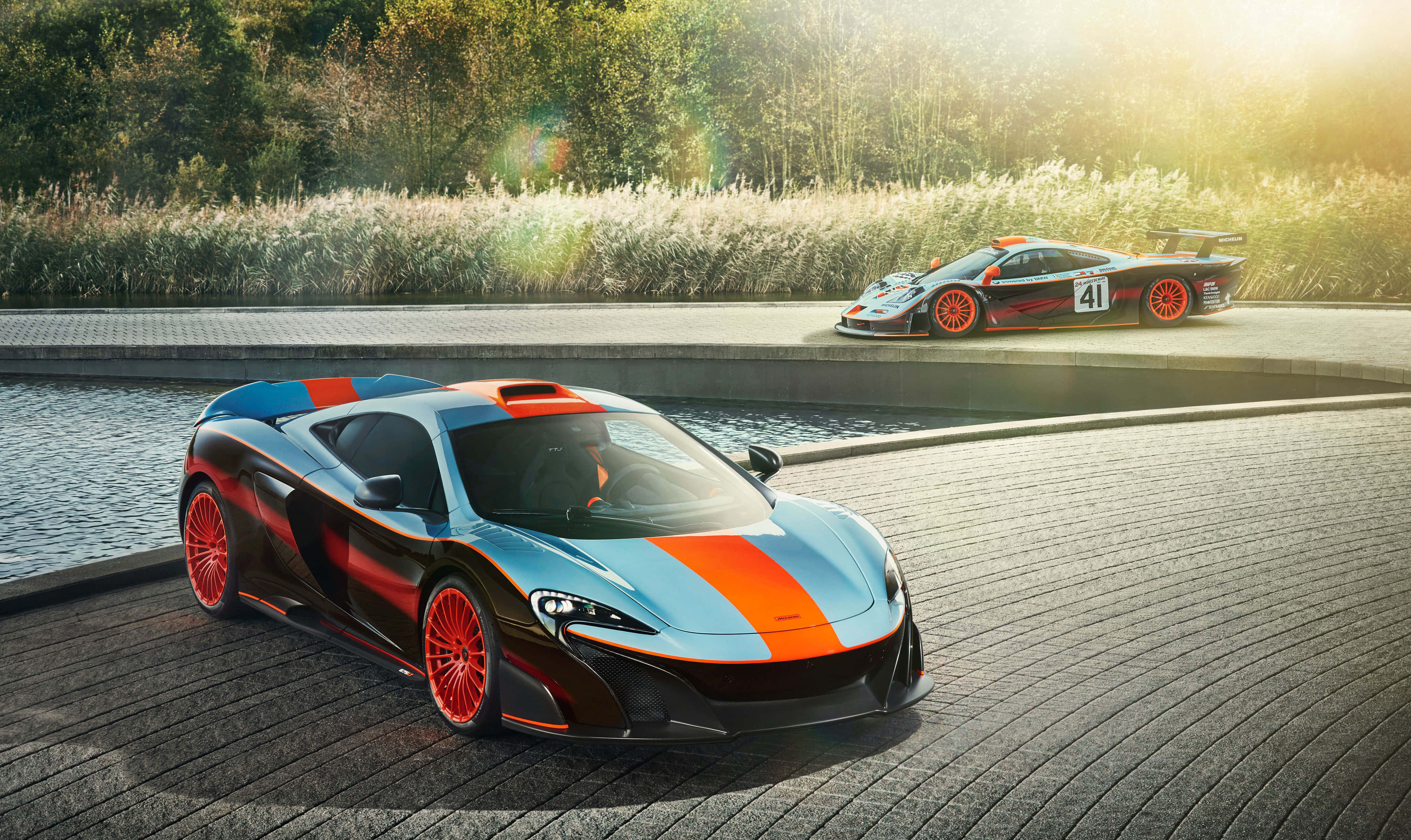 Mclaren F1 桌布 壁紙 — 兩輛時尚的McLaren跑車，帶有醒目的橙色點綴，在金色陽光下，在潮濕的鵝卵石小徑上閃閃發光，旁邊是反射水景。