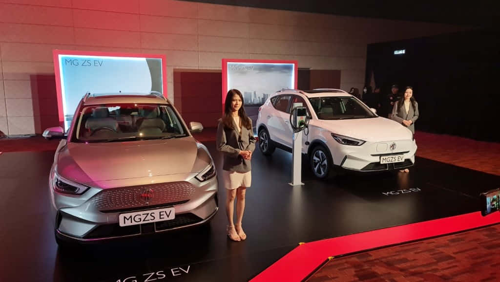 Mg Zs 桌布 壁紙 — 兩輛時尚的MG ZS EV電動車在紅地毯舞台上，主持人在旁，在戲劇性燈光下展示。