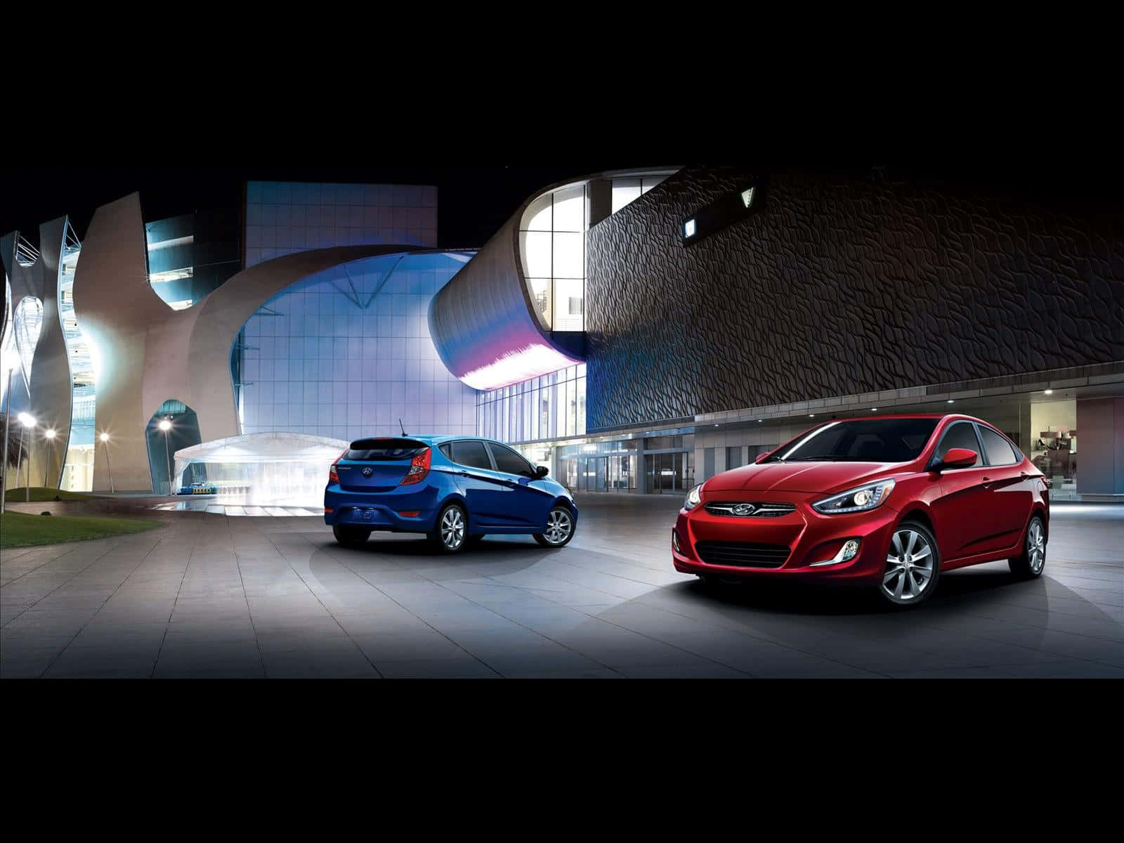 Hyundai Accent 桌布 壁紙 — 兩輛時尚現代汽車，一輛藍色掀背車和一輛紅色轎車，夜間停在一棟未來主義照明建築前。