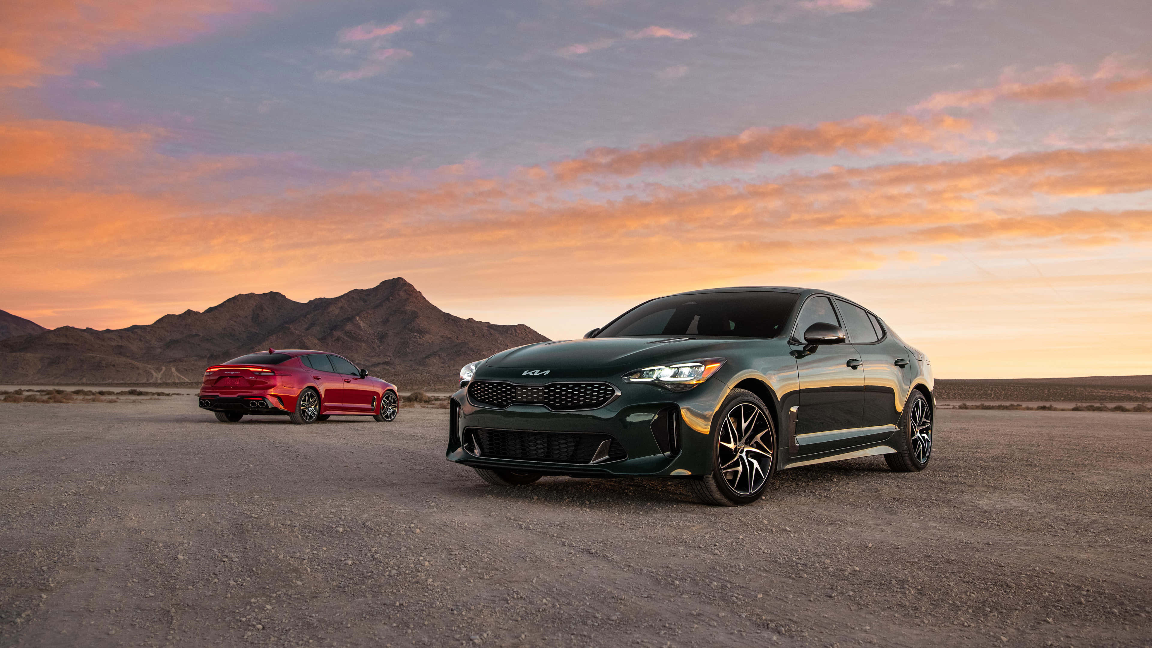 Kia Stinger 桌布 壁紙 — 兩輛時尚跑車，一紅一綠，停在沙漠碎石上，背景是 vibrant 的日落天空和山脈。