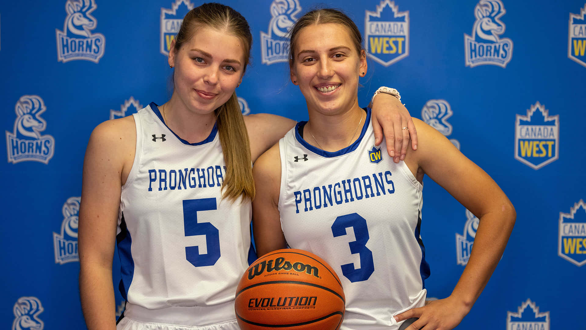  壁紙 — 兩名微笑的女籃球員身穿白色「PRONGHORNS」球衣，在藍色品牌背景前與Wilson籃球合影。