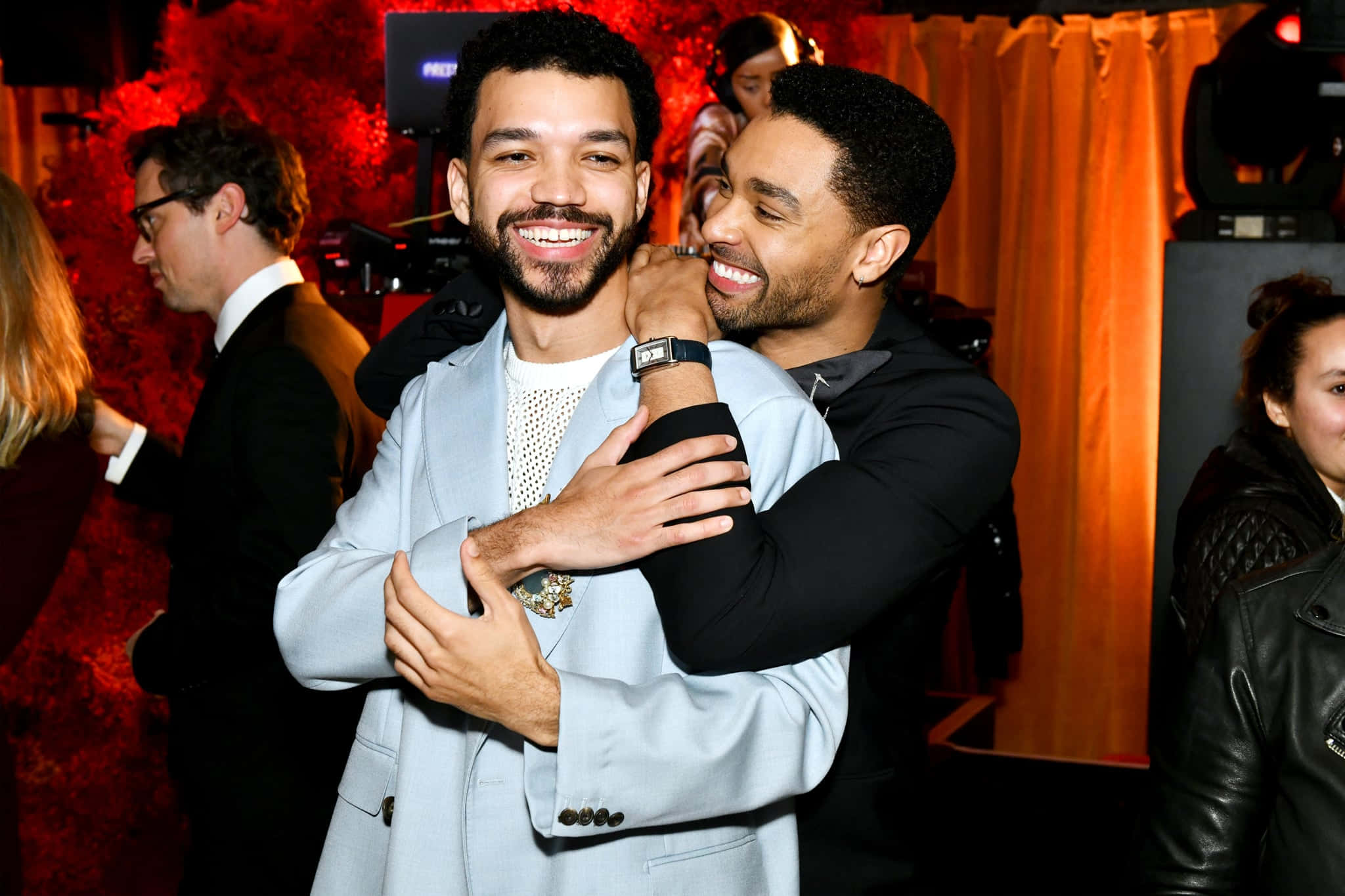 Justice Smith 桌布 壁紙 — 兩位微笑的男子在一個充滿活力的紅色燈光活動中熱情擁抱，背景有橙色窗簾和其他賓客。