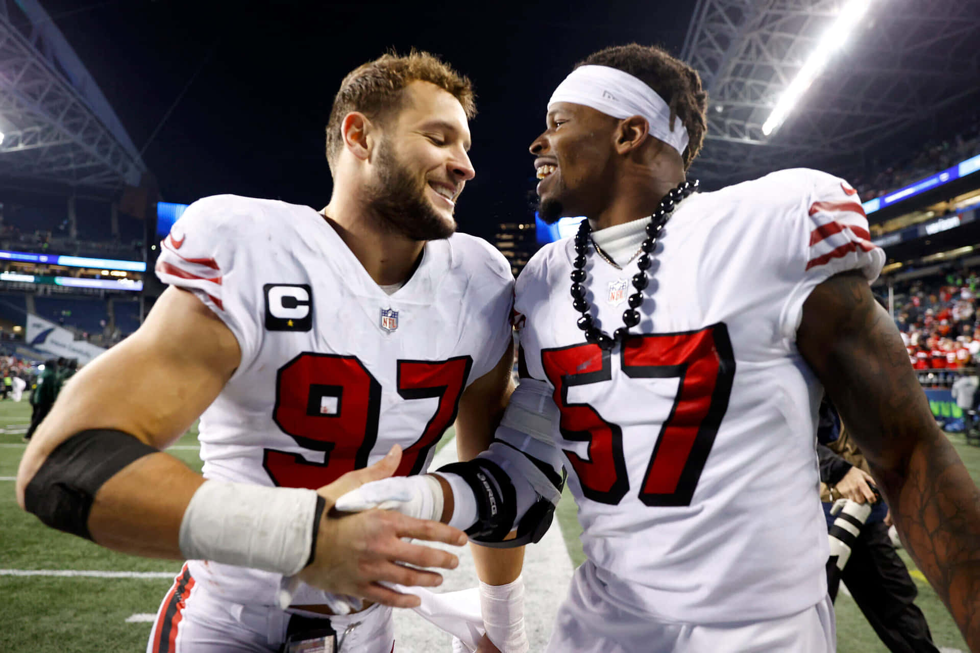  壁紙 — 兩名身穿白色球衣、紅色號碼97和57的NFL球員面帶微笑，在夜間燈光明亮的足球場上愉快地握手。