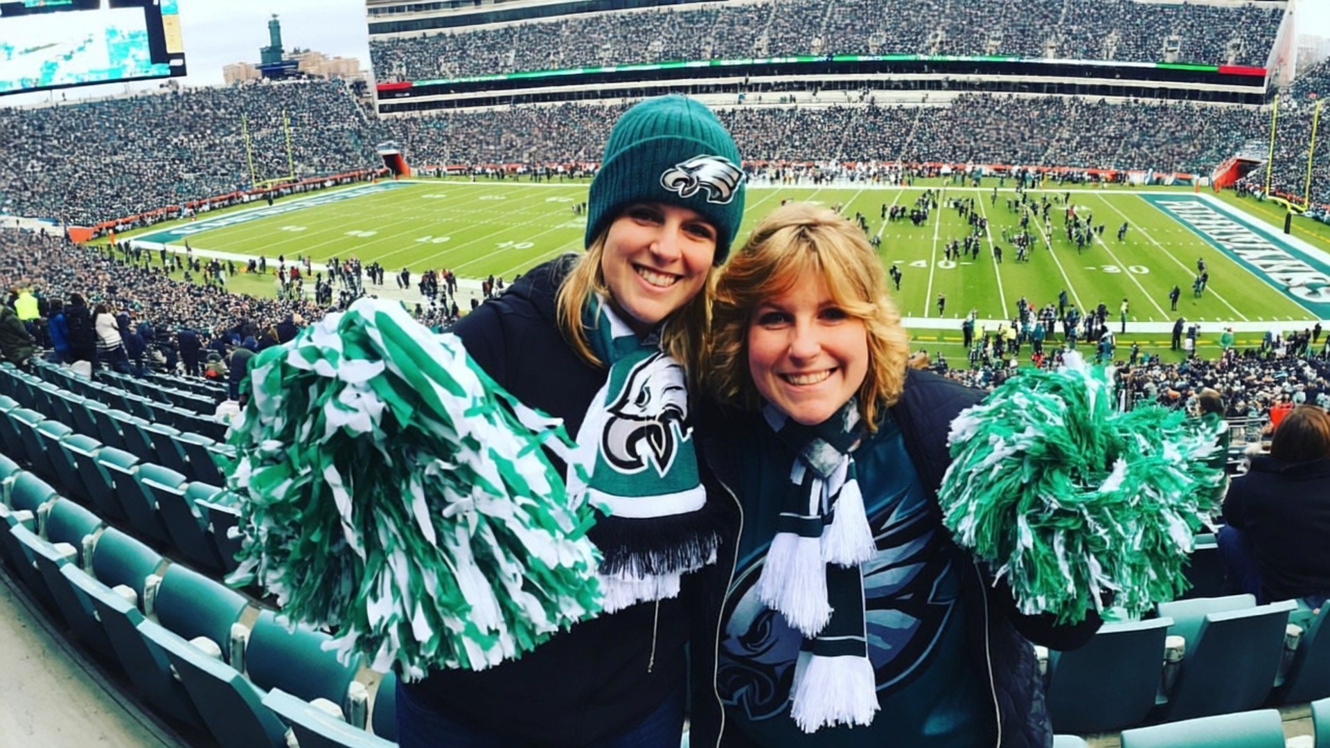 Eagles Fans Celebrating 壁紙 — 兩位面帶微笑、身穿老鷹隊裝備的女性在擠滿人的足球場上揮舞著綠色和白色的絨球。