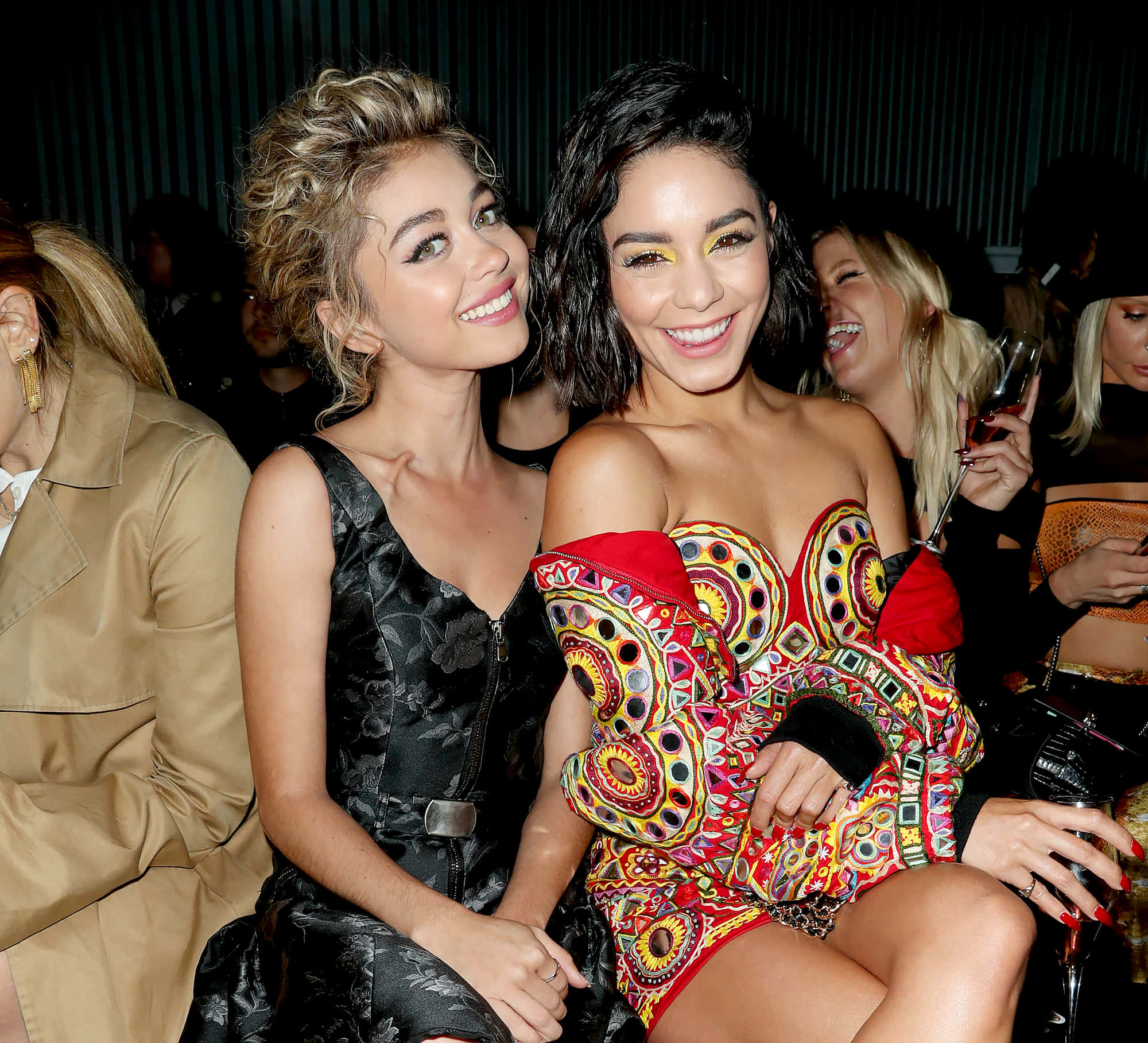 Sarah Hyland 桌布 壁紙 — 兩位微笑的女性穿著華麗服裝，在充滿活力、色彩繽紛圖案和溫暖燈光的社交活動中一起擺姿勢。