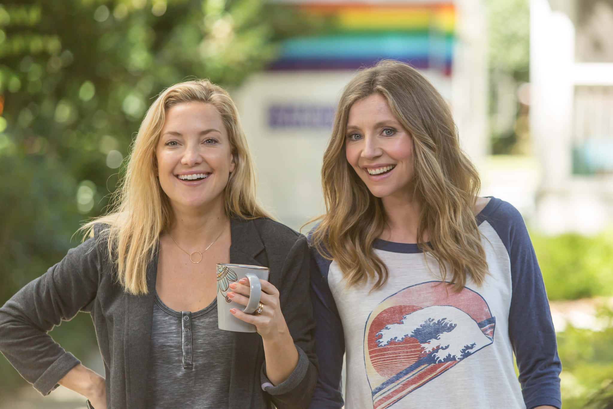 Sarah Chalke 桌布 壁紙 — 兩位微笑的女性站在戶外，一位拿著馬克杯，另一位穿著波浪圖案T恤，身後可見彩虹旗。