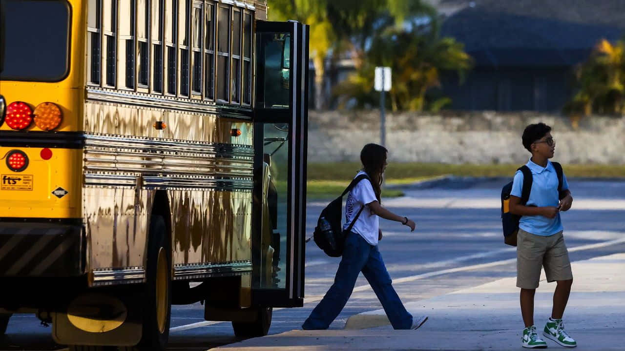  壁紙 — 兩名背著背包的學生在陽光明媚的街道上走過一輛黃色校車，背景有棕櫚樹。