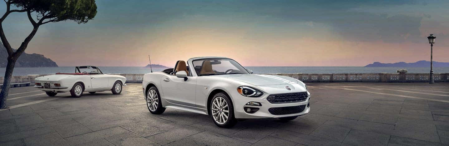 Fiat 124 Spider 桌布 壁紙 — 兩輛白色敞篷跑車停泊在石頭步道上，俯瞰日落時平靜的大海。
