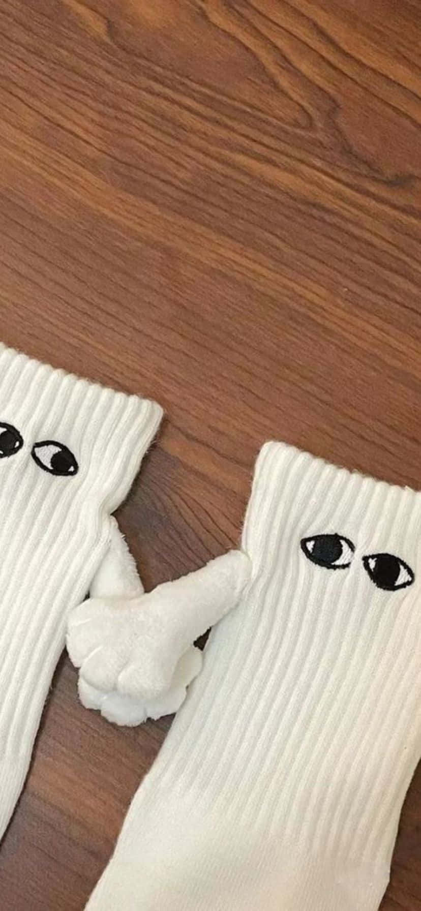  壁紙 — 兩隻白色羅紋襪，帶有黑色卡通眼睛，一隻白色毛絨小手在木地板上豎起一根手指。