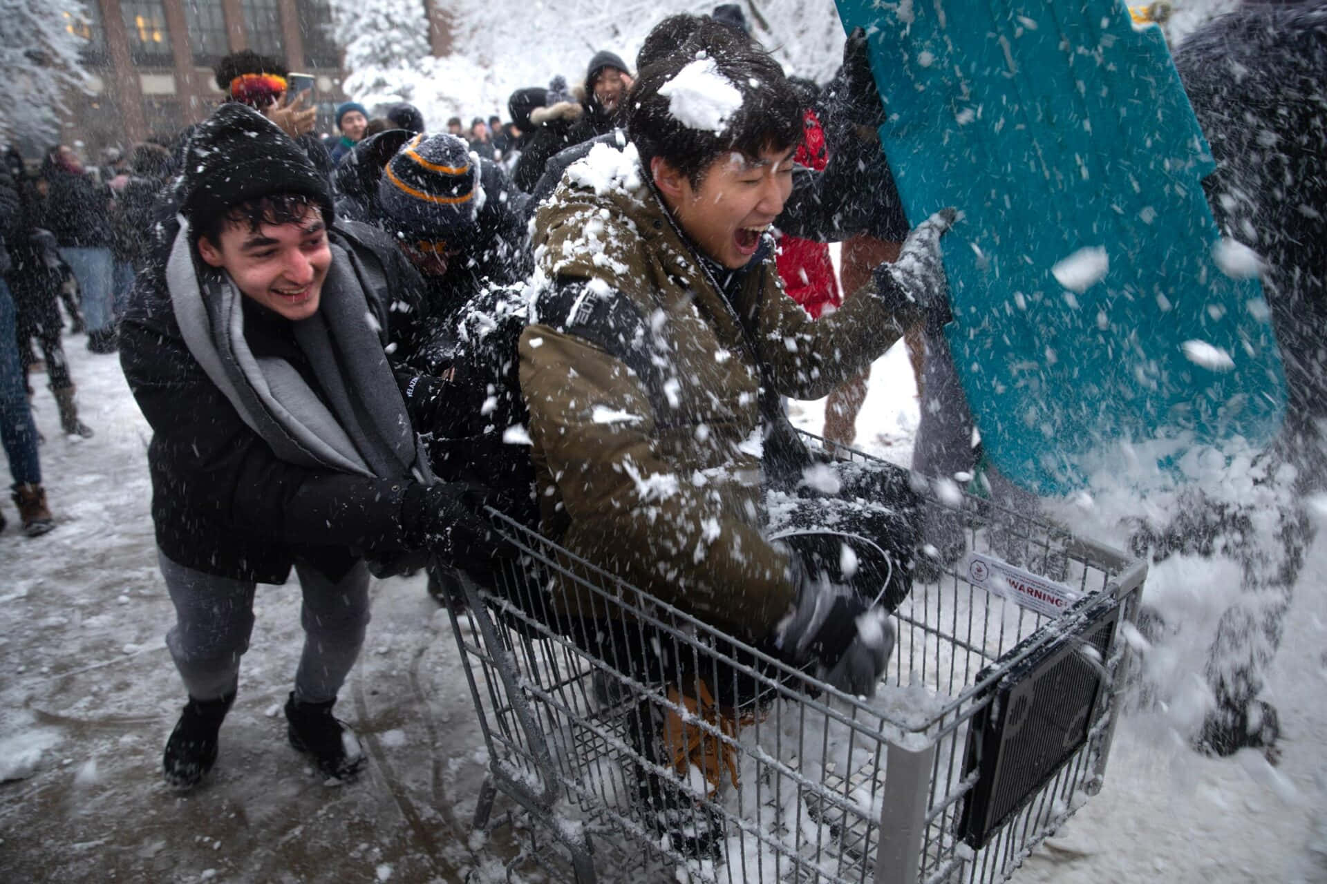  壁紙 — 兩個年輕男子笑著推著裝滿雪的購物車穿過積雪的城市街道。