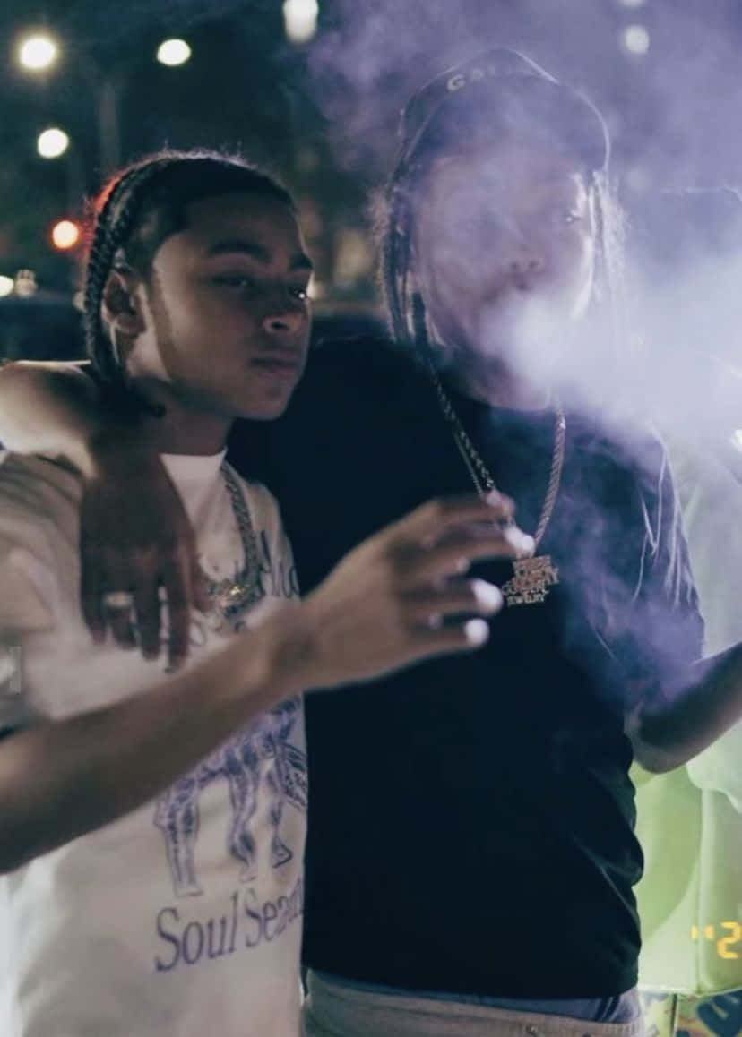  壁紙 — 兩個年輕男子在夜晚靠近站立，一人呼出濃煙，另一人在城市燈光下向前看。