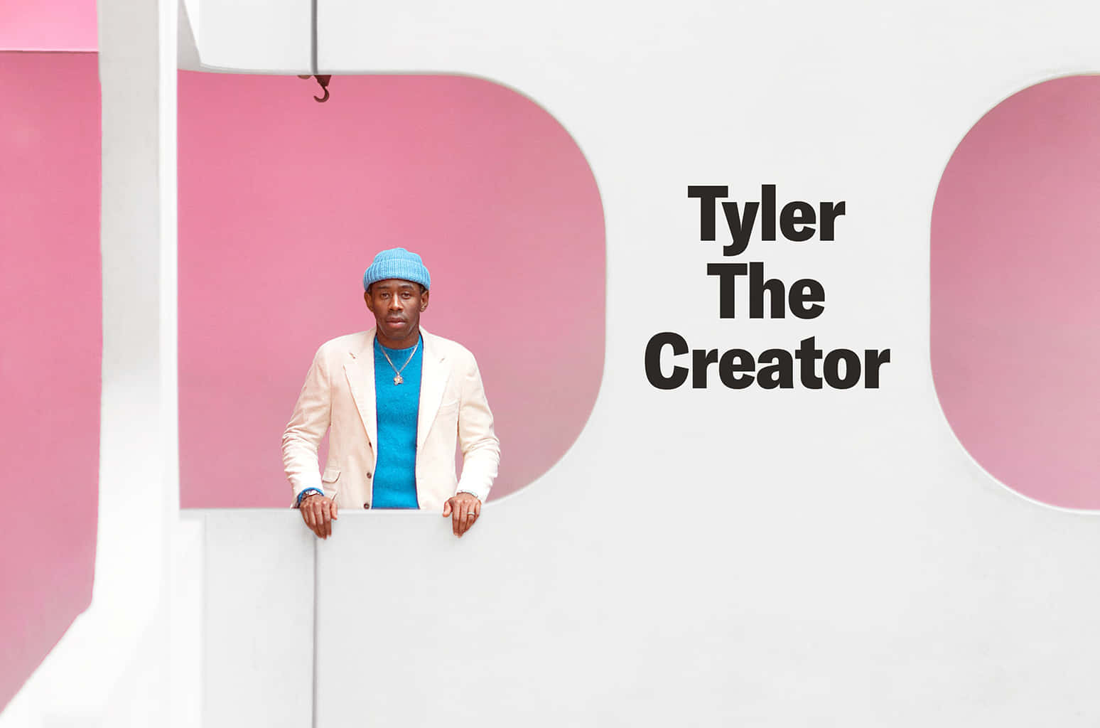 造物主泰勒 桌布 壁紙 — Tyler The Creator 靠在一個白色邊緣上，面對一個大膽的粉紅和白色幾何牆，牆上以黑色字體寫著他的名字。