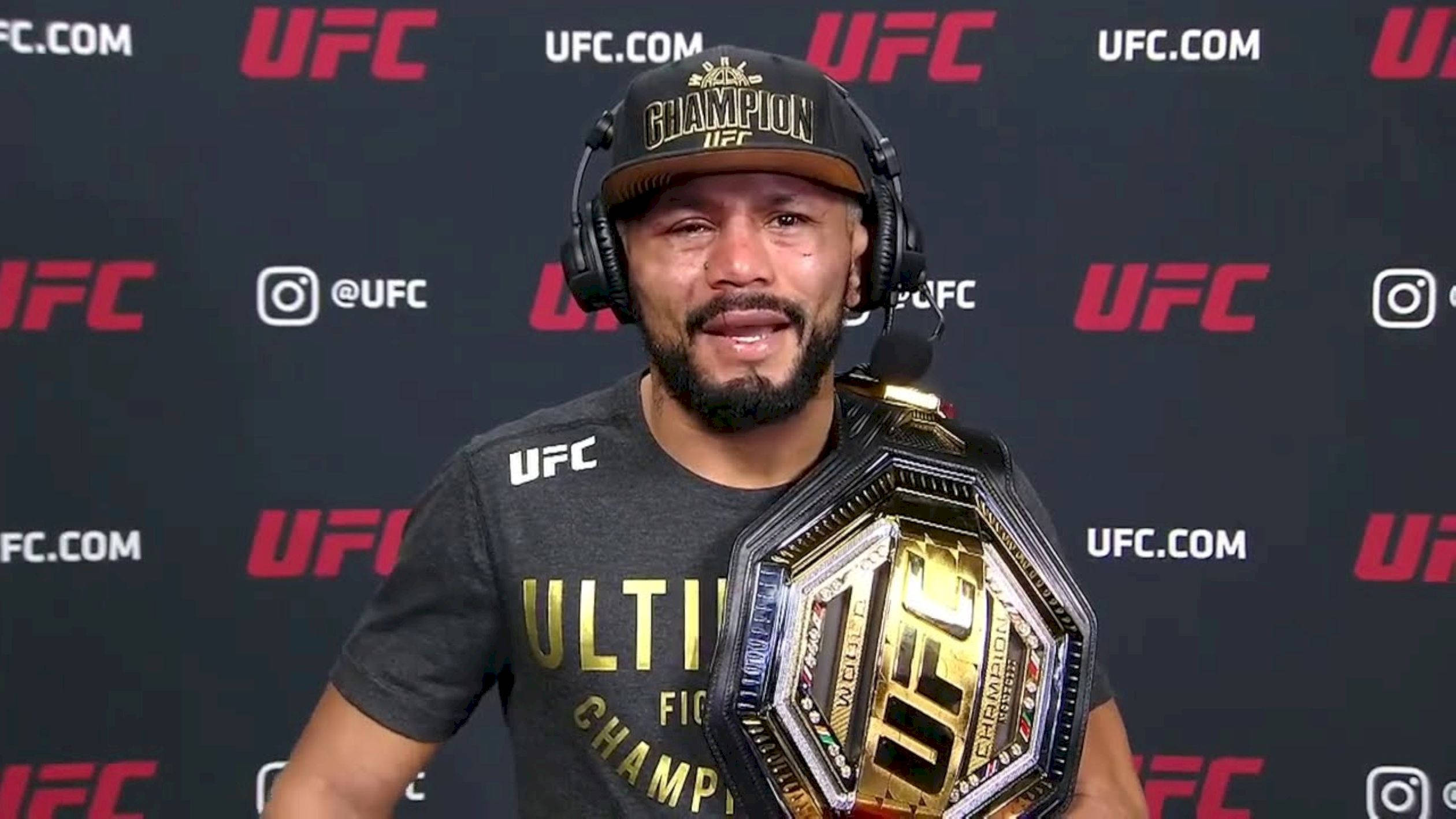 UFC 桌布 壁紙 — UFC冠軍拳手自豪地舉起冠軍腰帶，戴著「Champion」帽和耳機，背景為品牌背景。