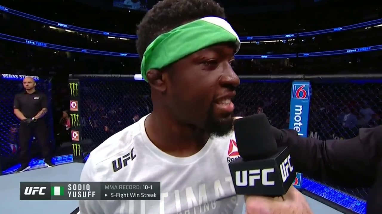 MMA 桌布 壁紙 — UFC拳手Sodiq Yusuff身穿白色裝備和綠色頭帶，在明亮的場館燈光下於八角籠內對著麥克風說話。