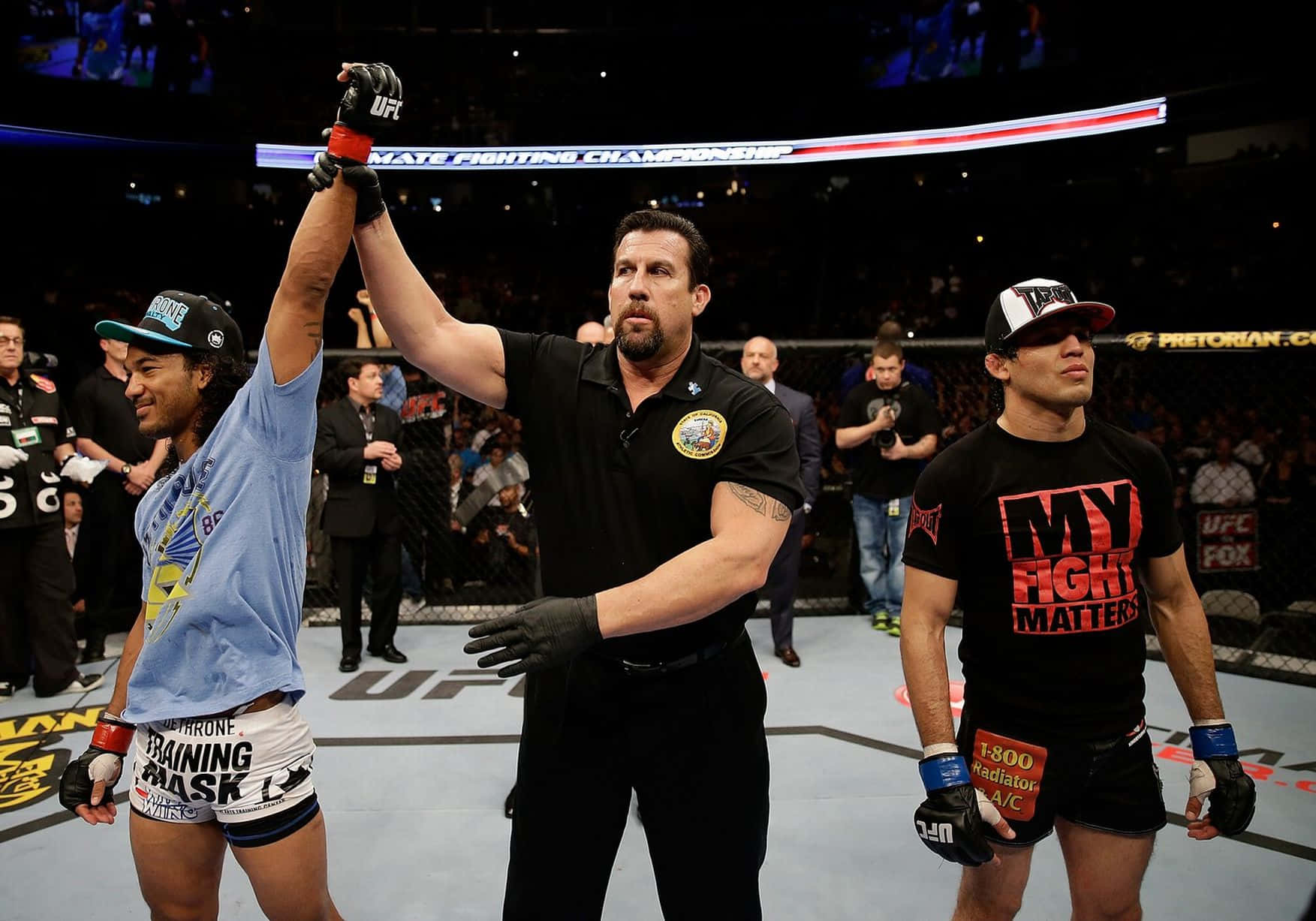 MMA 桌布 壁紙 — UFC裁判在八角籠中舉起勝利的拳手的手，而對手在場館燈光下沮喪地站著。