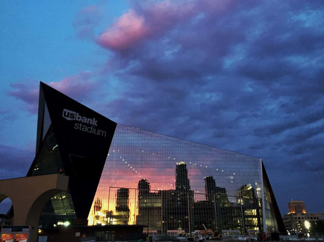 Bank 桌布 壁紙 — US Bank Stadium 的玻璃外牆映照出充滿活力的城市天際線，背景是帶有粉紅色和紫色雲彩的暮色天空。