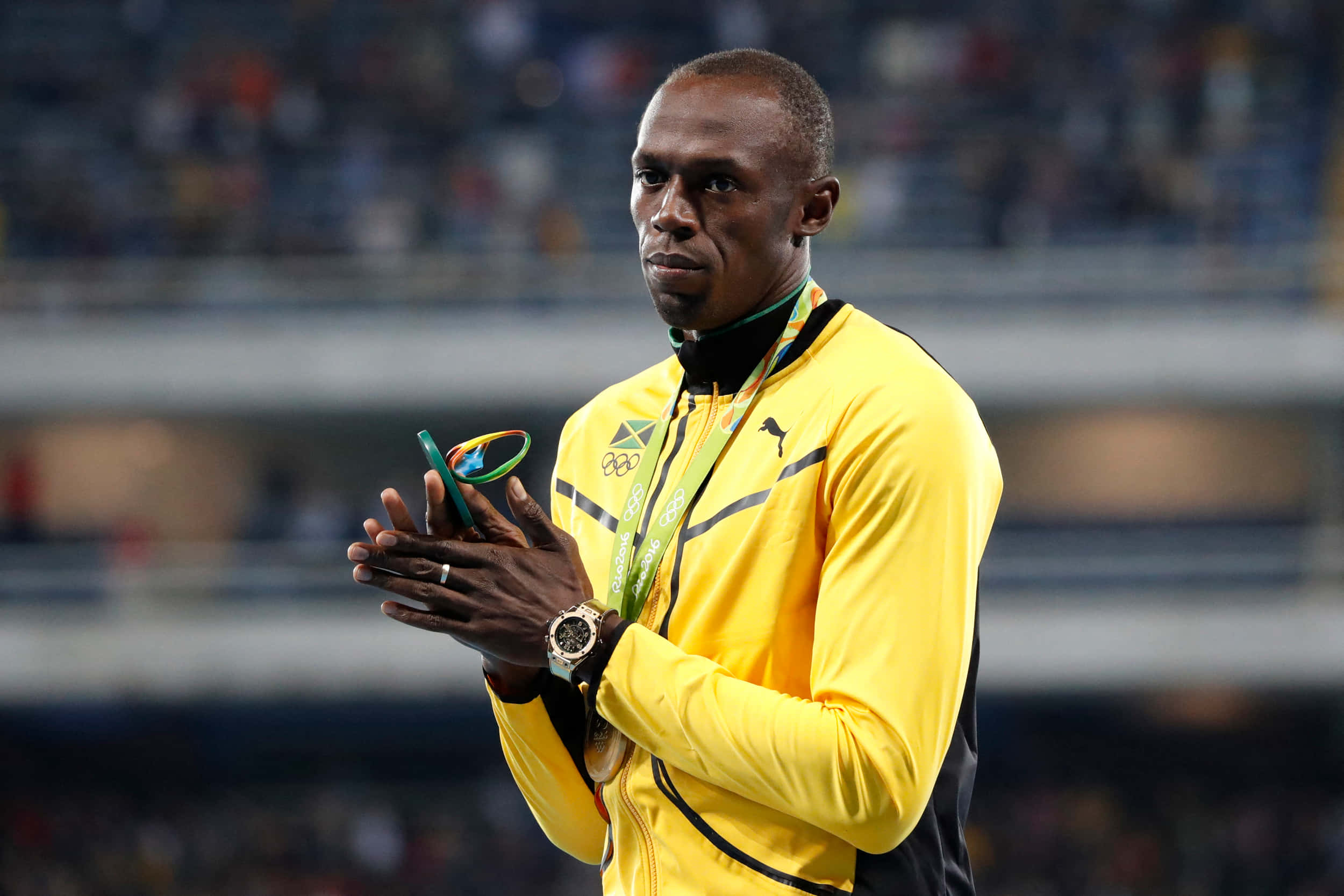 運動 桌布 壁紙 — Usain Bolt 身穿黃色牙買加運動服，在明亮燈光下的體育館中手持銅牌鼓掌。