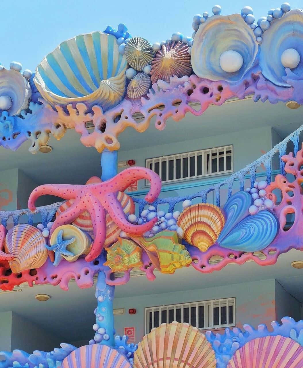  壁紙 — 色彩繽紛的3D海洋生物雕塑，包括貝殼、章魚和珊瑚，在晴朗的藍天下裝飾著建築物的外牆。